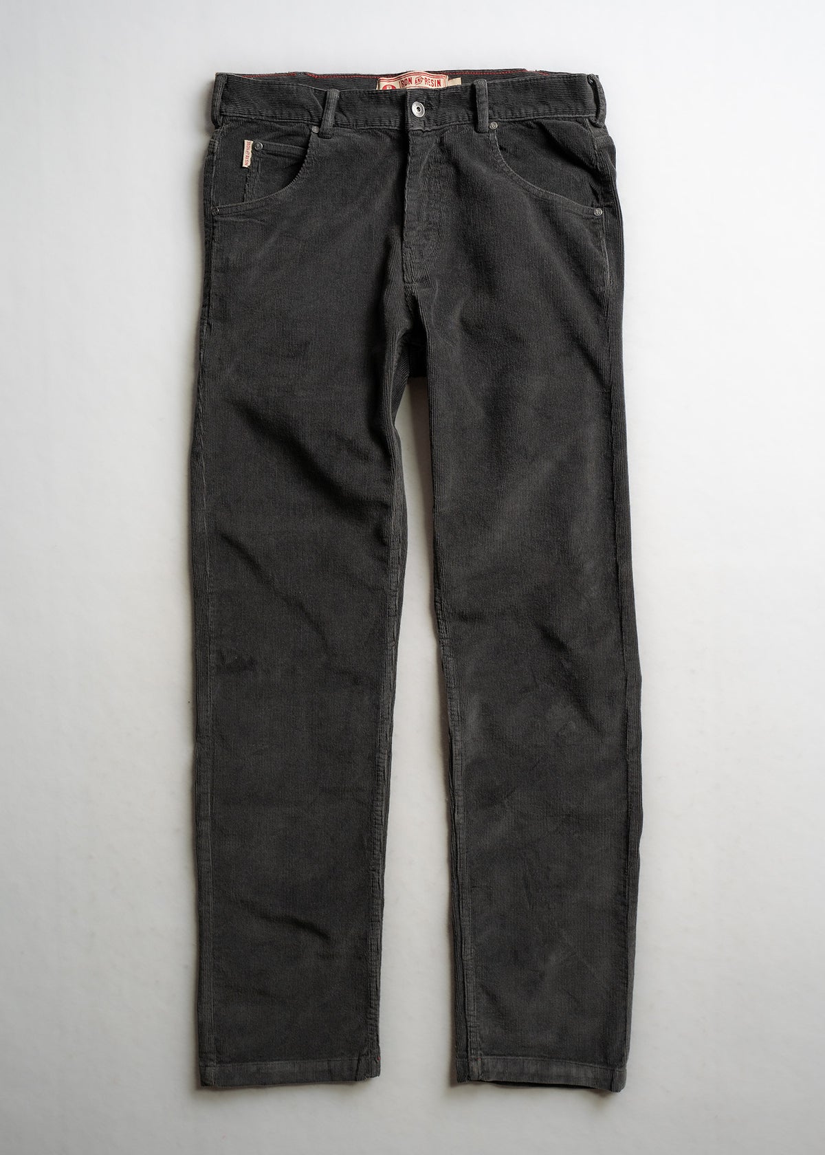 Dark gray corduroy pants with front pockets and button closure, laid flat on a light background, showcasing texture and design.