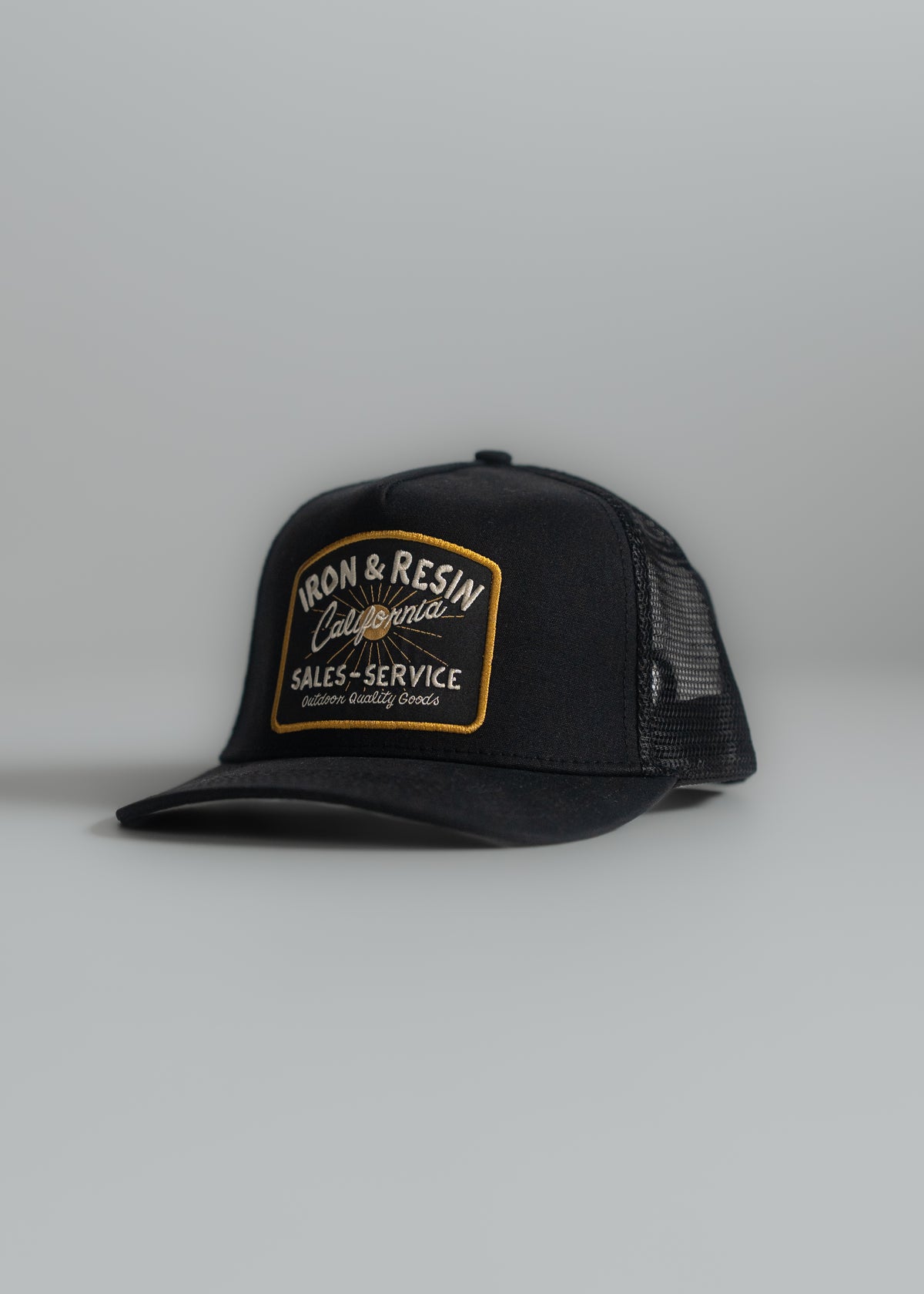 Black trucker hat with "Iron & Resin California" patch, featuring gold embroidery, on a light gray background.