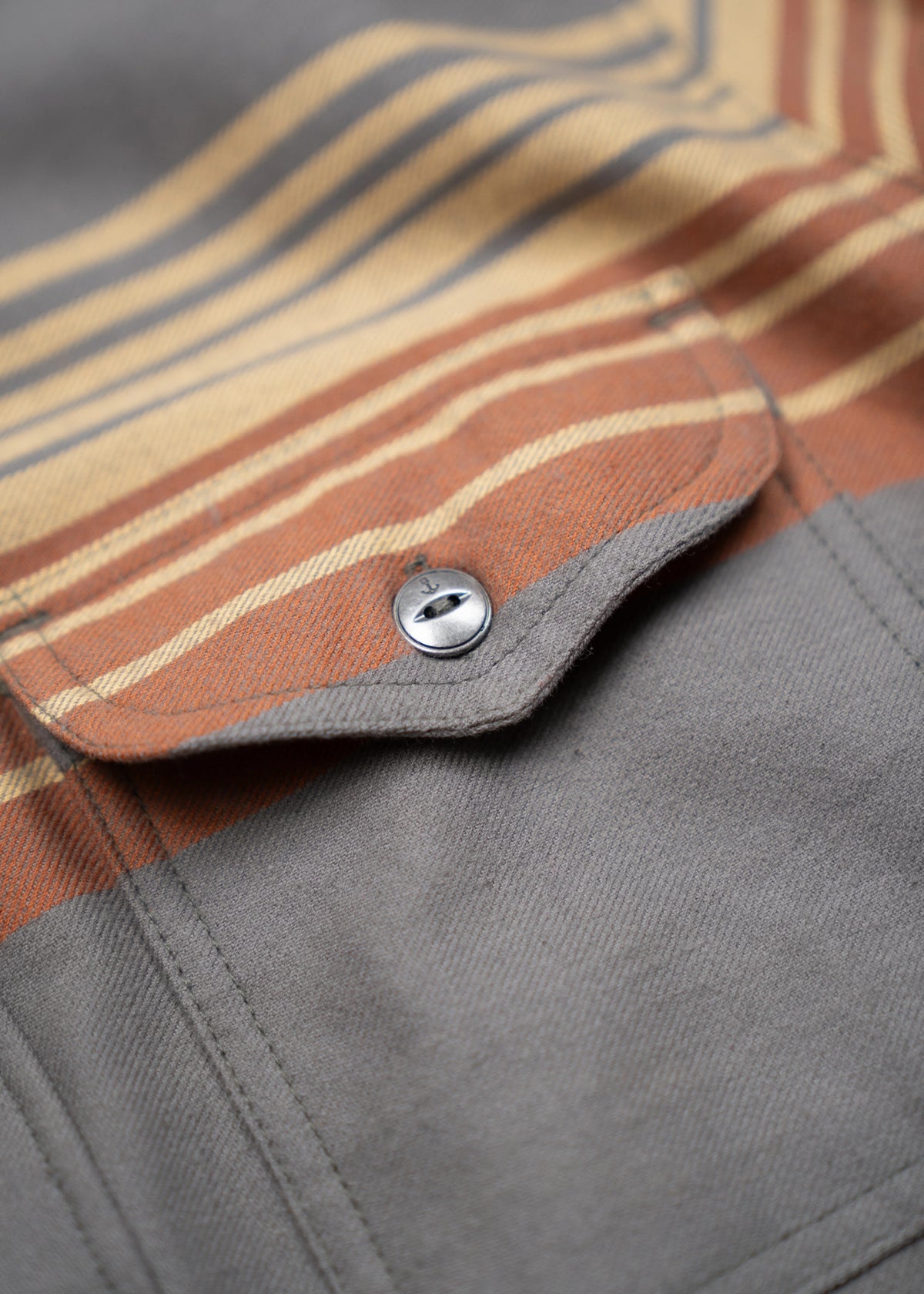 Close-up of a gray shirt pocket with a button, featuring orange and beige stripes above, creating a stylish, casual look.