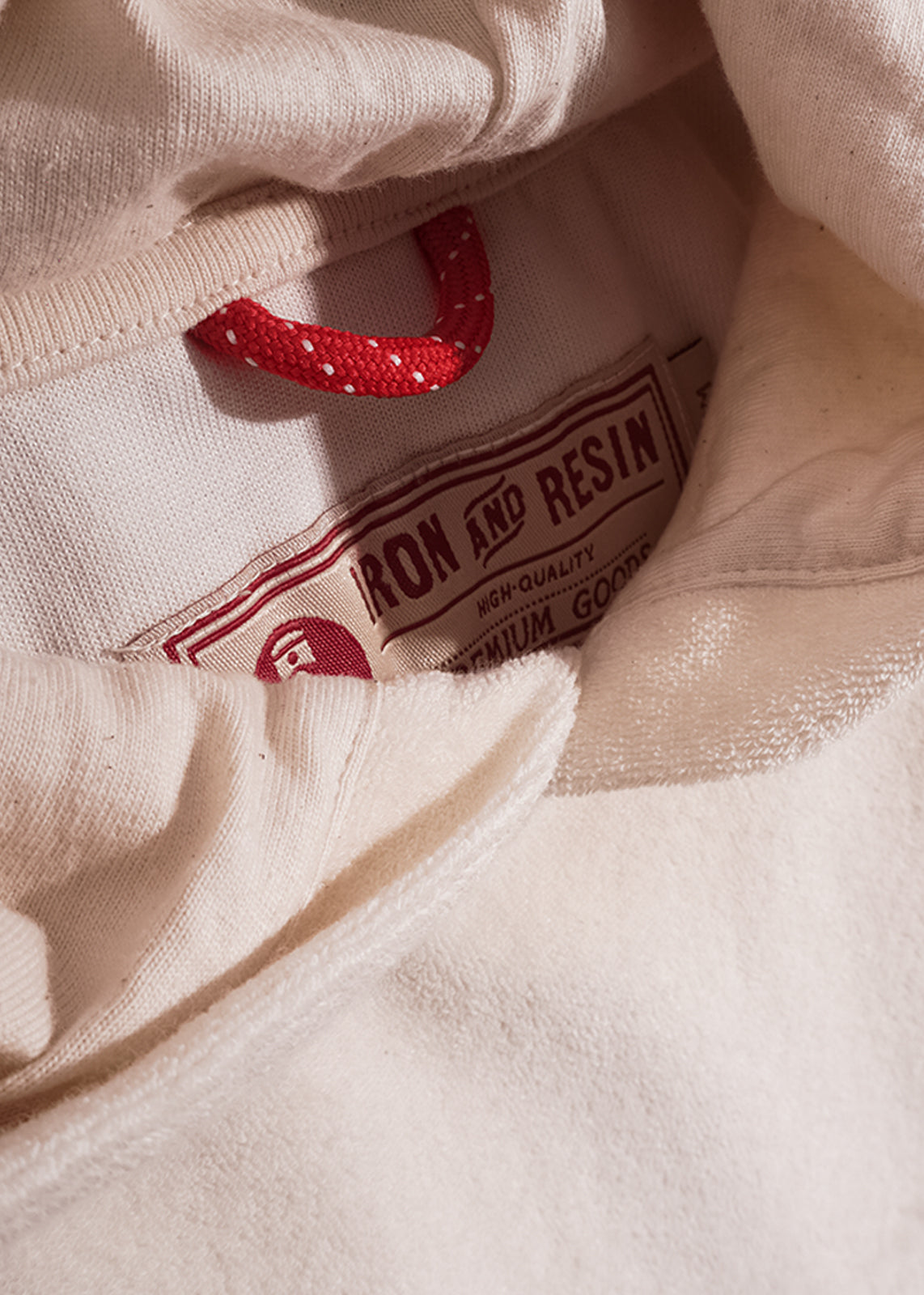Close-up of a cream hoodie with a red drawstring and "Iron and Resin" label, highlighting its high-quality, premium design.