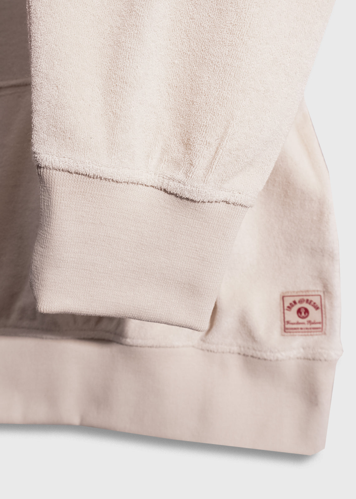 Close-up of a beige sweatshirt sleeve with ribbed cuff and a small red label on the hem, displaying texture and stitching details.