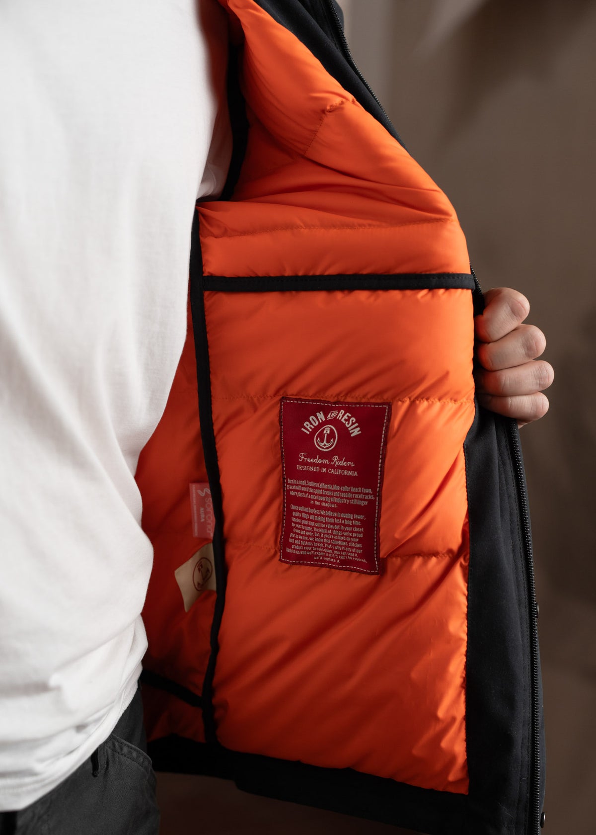Man in white shirt holds open black jacket, revealing orange lining with a red label reading "Iron & Resin Freedom Riders."
