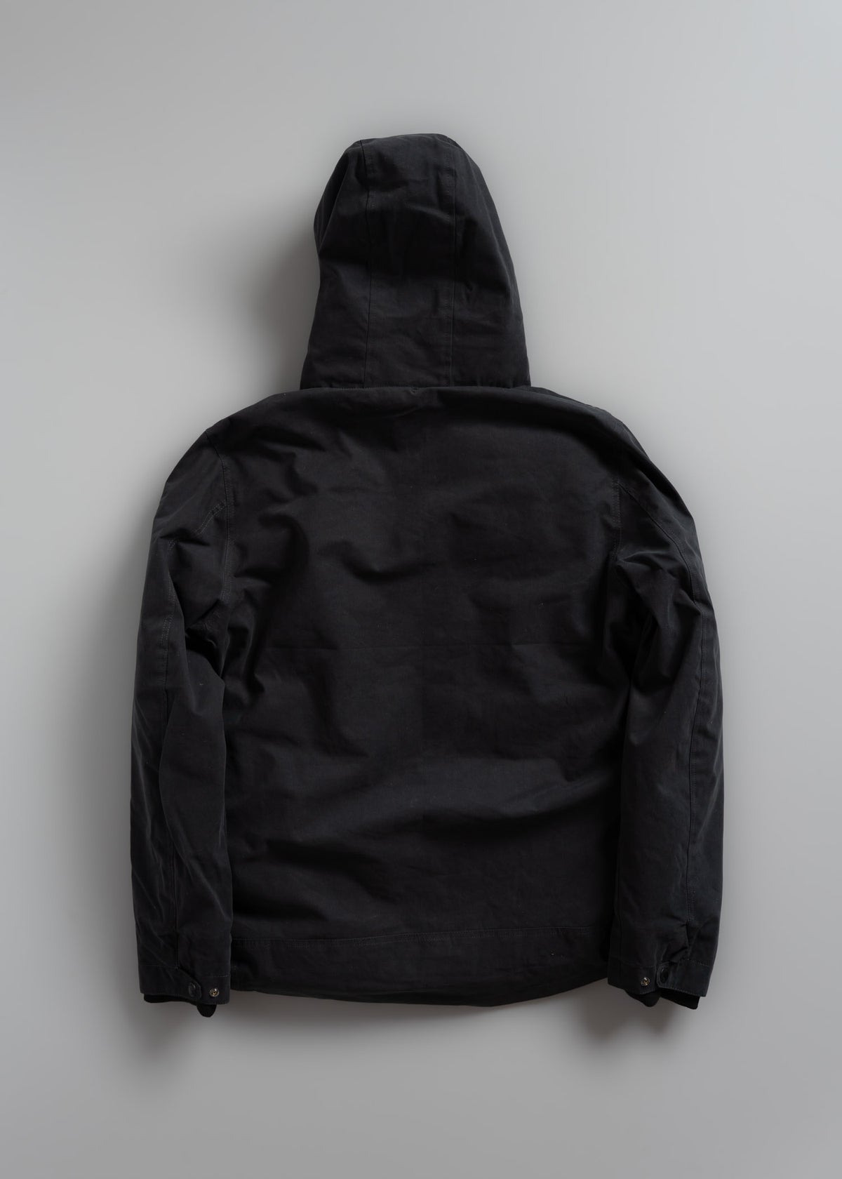 Black hooded jacket laid flat on a light gray background, showing the back view with long sleeves and a relaxed fit.