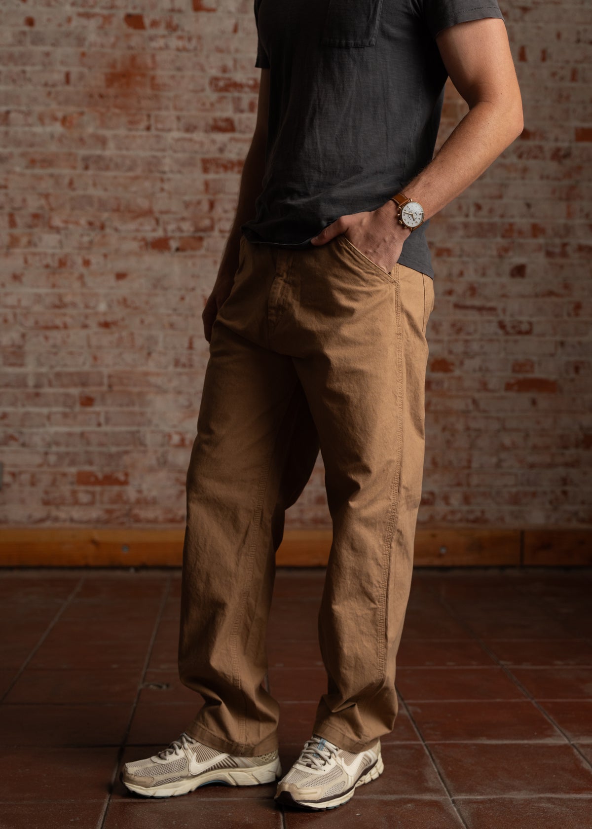 Man in casual outfit with brown pants and gray shirt, wearing a watch, stands against a brick wall, hands in pockets.