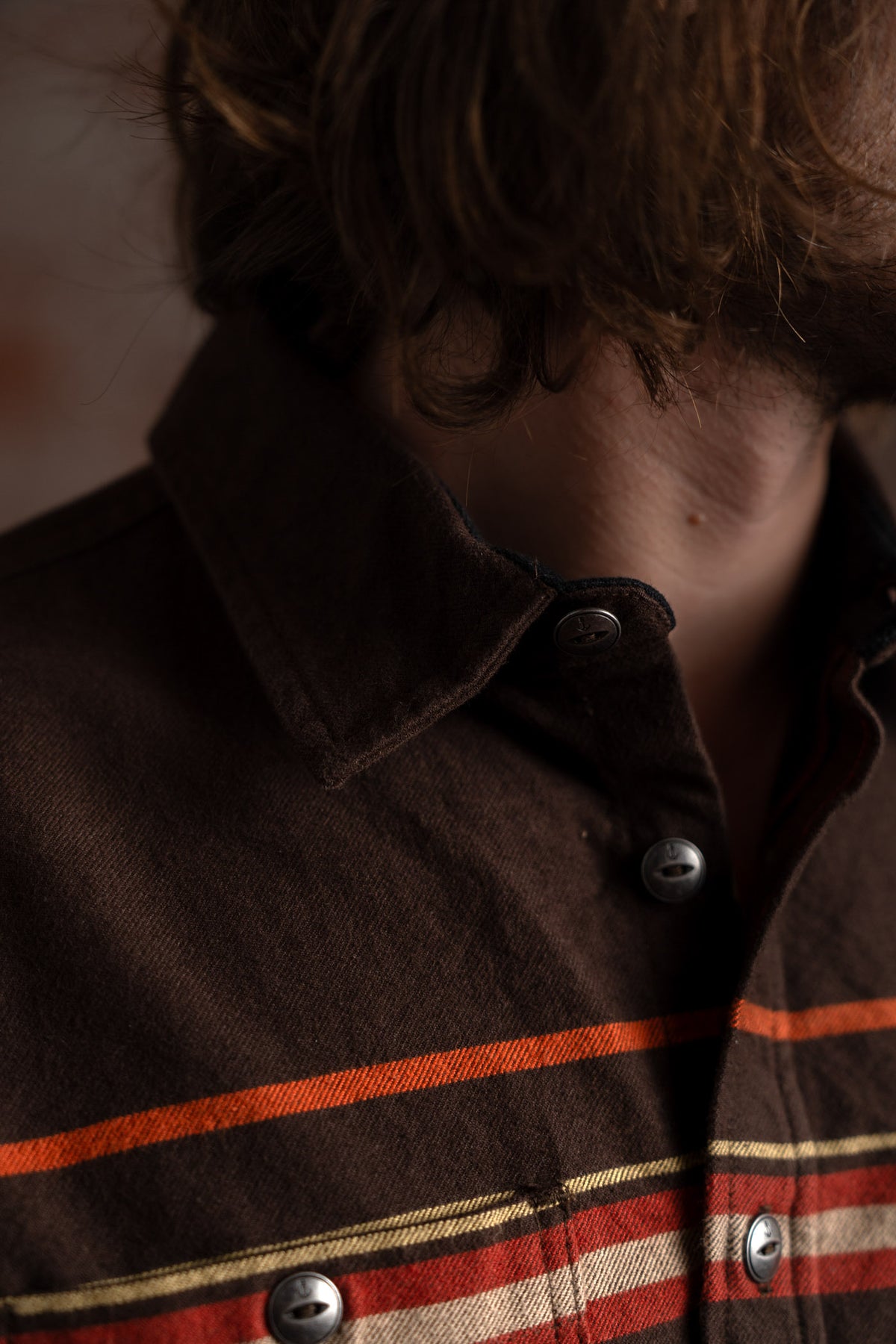 Close-up of a person wearing a brown shirt with red and orange stripes, highlighting the collar and buttons, hair slightly visible.