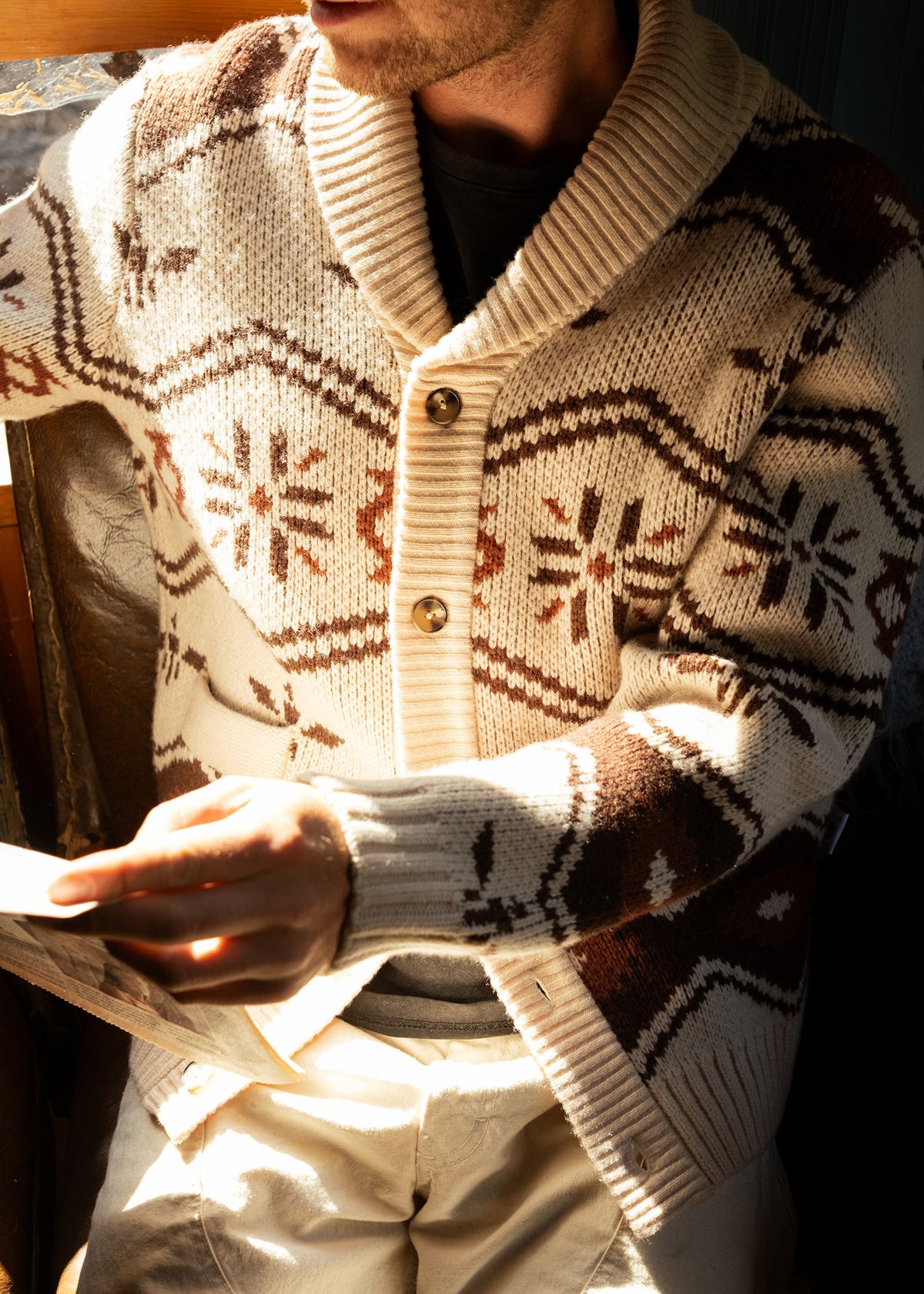 Man in a patterned cardigan reads a book by sunlight, highlighting the cozy knit texture and warm tones of the sweater.