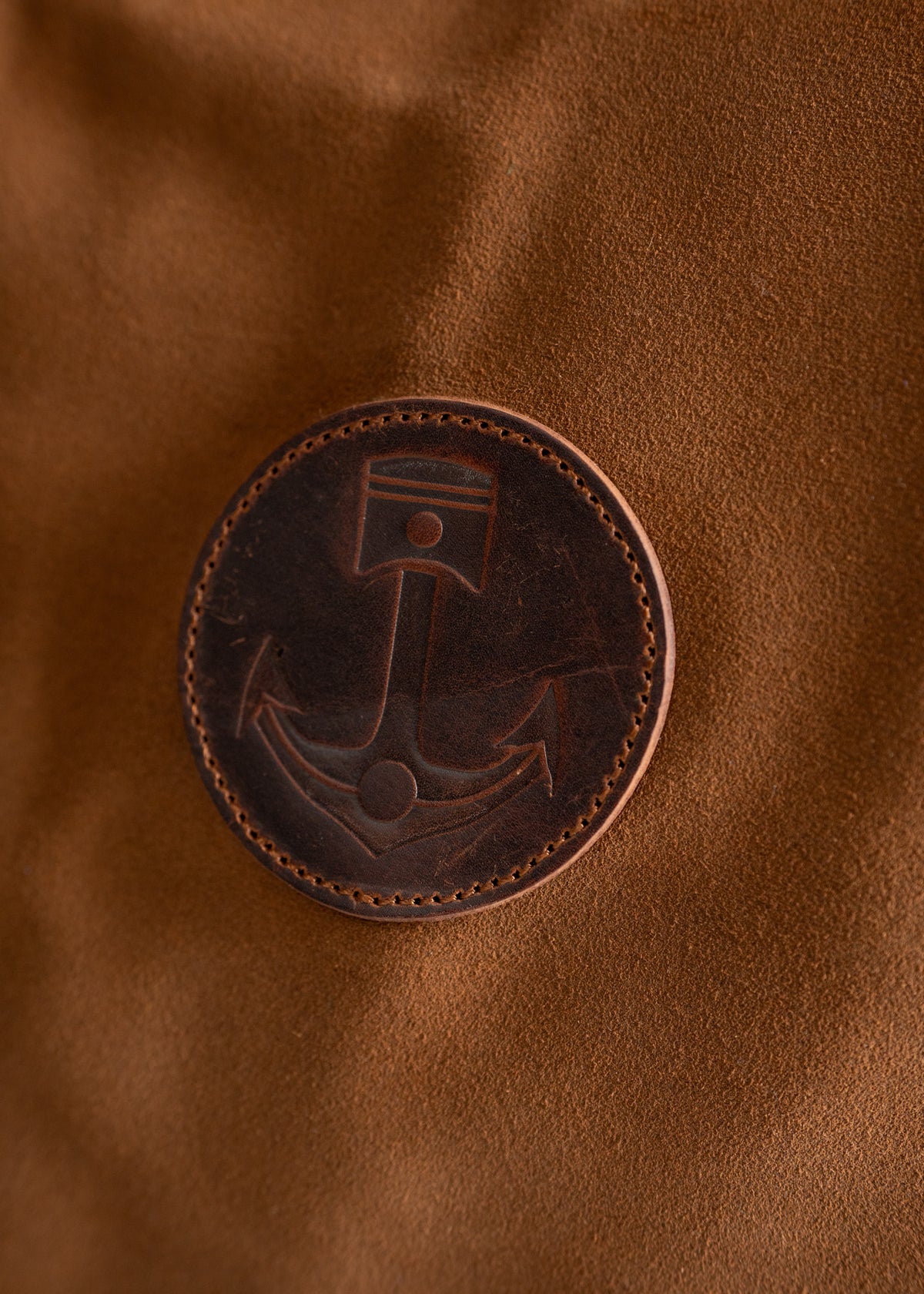A brown leather patch with an anchor design is stitched onto a textured tan fabric, creating a nautical-themed accessory.