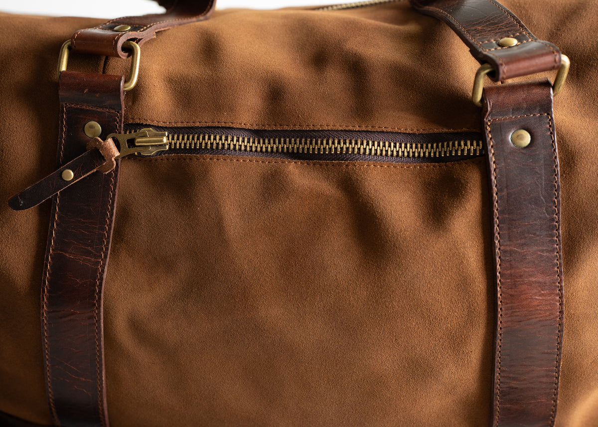Close-up of a brown leather duffel bag with a brass zipper and dark leather straps, showcasing craftsmanship and texture.
