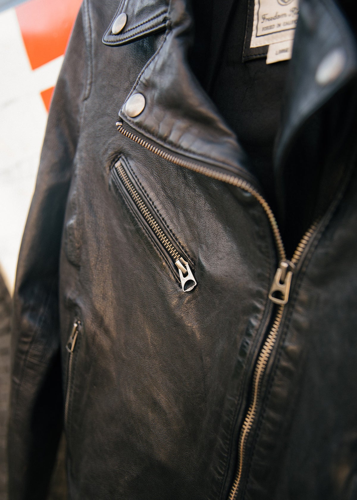 Close-up of a black leather jacket with silver zippers and buttons, hanging against a blurred red and white background.