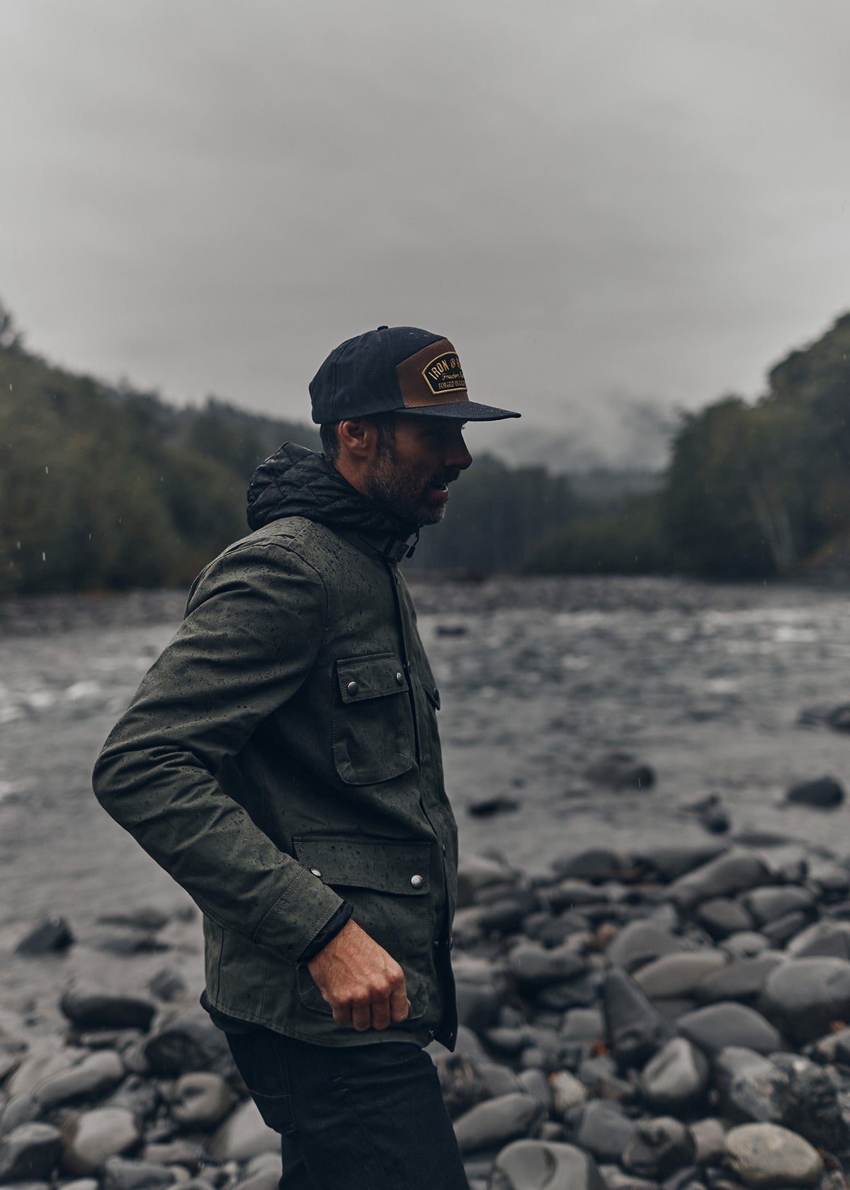 Man in a green jacket and cap stands by a rocky riverbank under a cloudy sky, surrounded by misty forest.