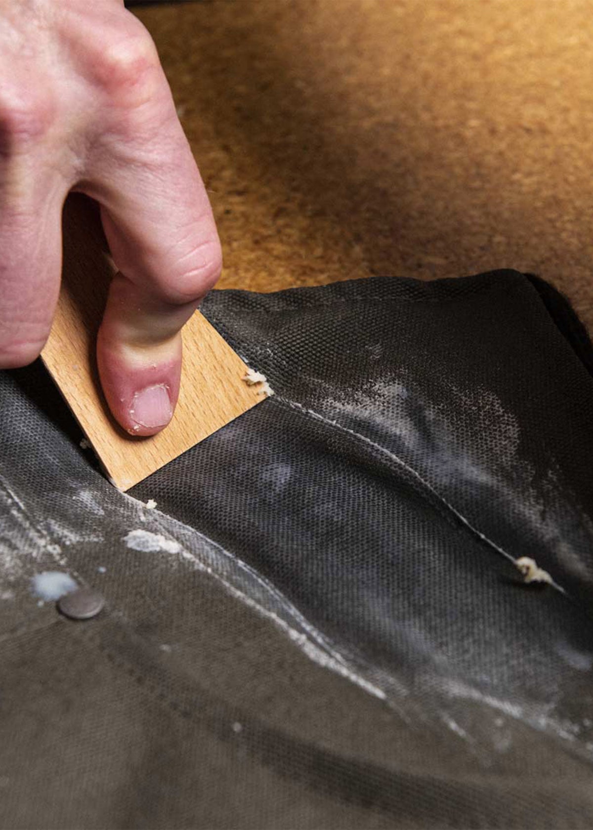 Hand scraping excess glue from black fabric with a wooden tool on a brown surface, focusing on precision and craftsmanship.
