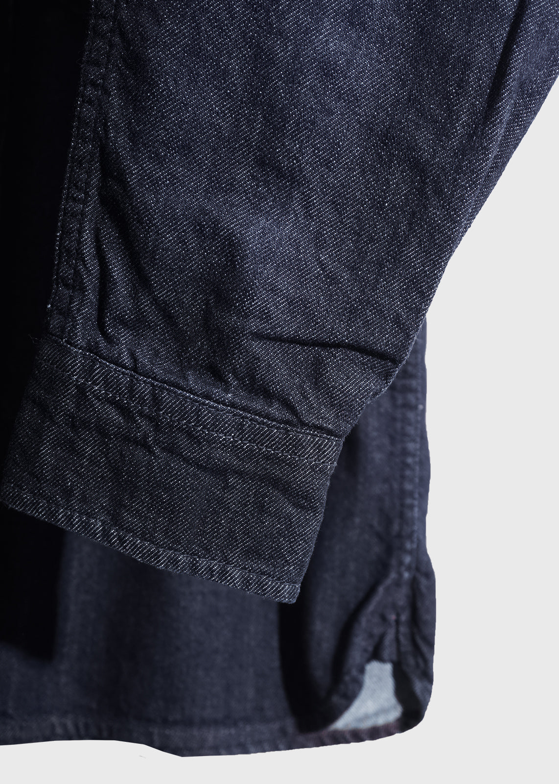 Close-up of a dark denim shirt sleeve with visible stitching, highlighting texture and fabric detail against a light background.