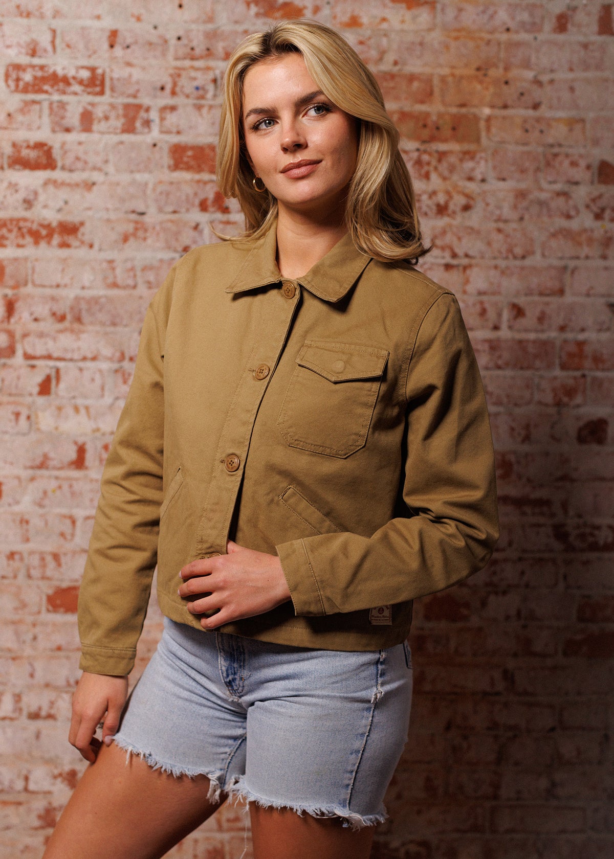 Woman in tan jacket and denim shorts stands against a brick wall, looking to the side with a thoughtful expression.