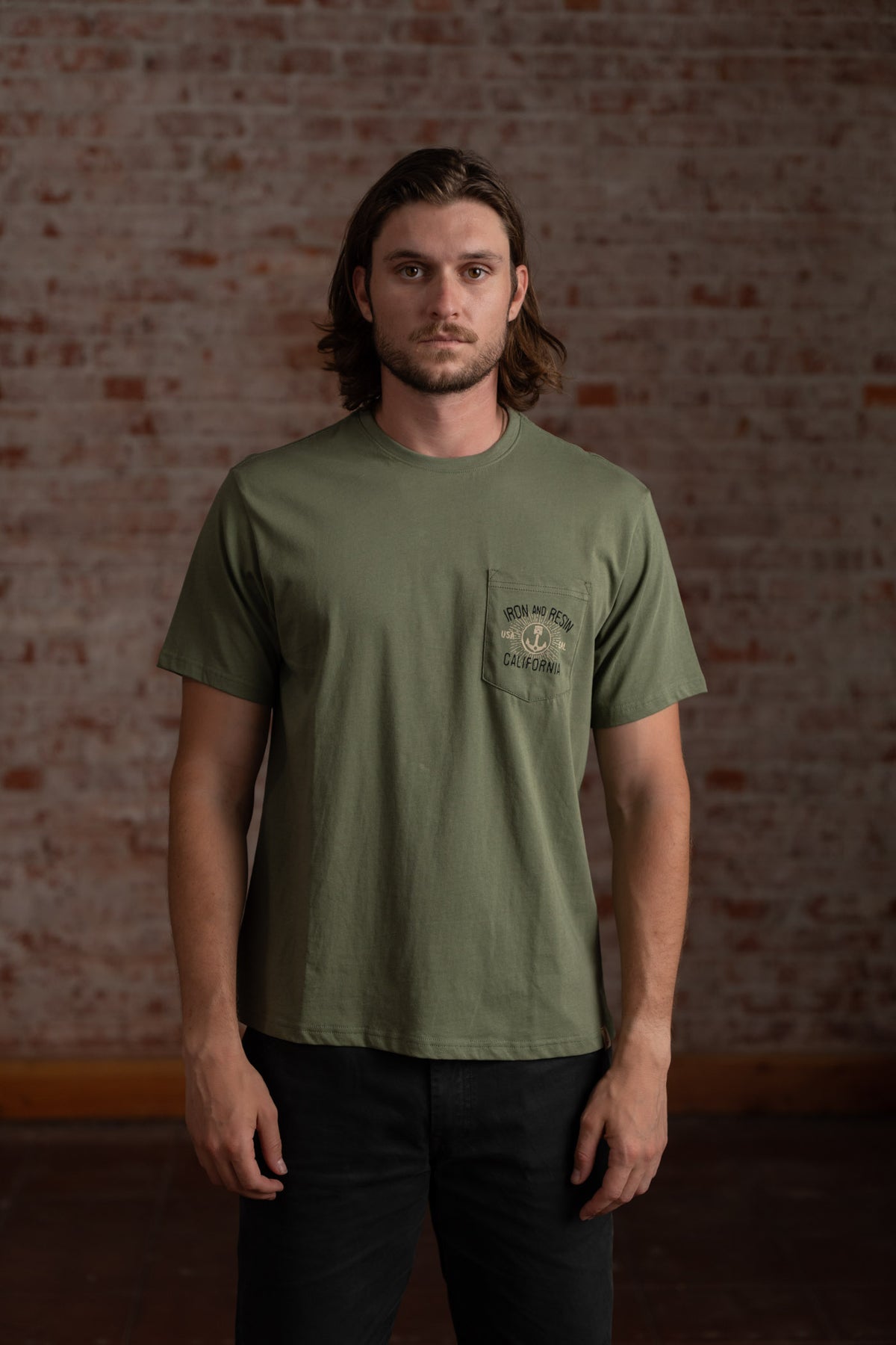 Man in olive green t-shirt stands against a brick wall, exuding a calm demeanor. Text on shirt reads "Iron and Resin California."