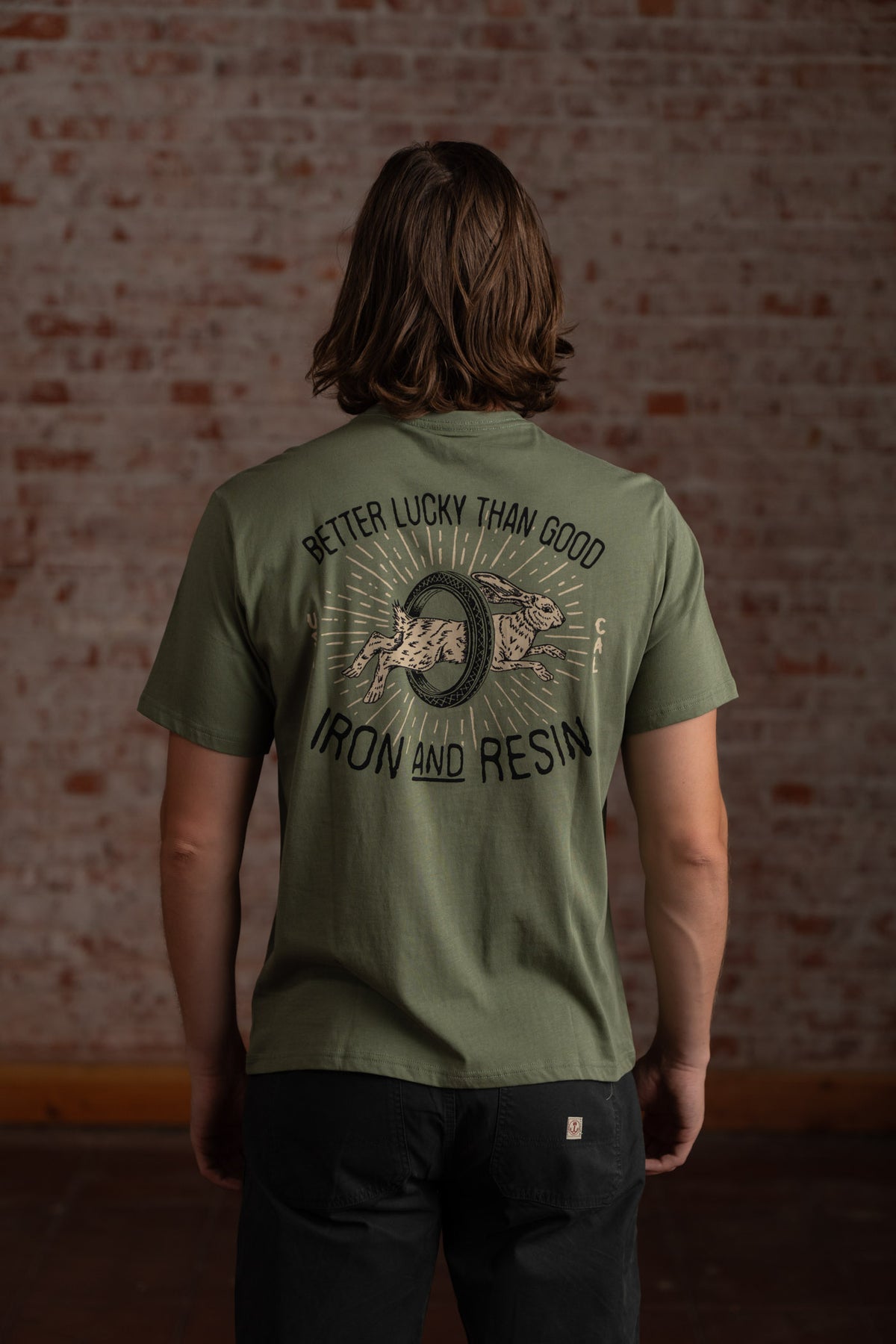 Man in green "Better Lucky Than Good" Iron and Resin shirt stands against a brick wall, showcasing the back design.