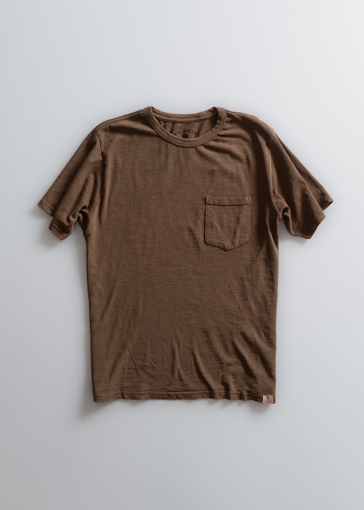 Brown T-shirt with a chest pocket, laid flat on a light background, showcasing its simple design and casual style.