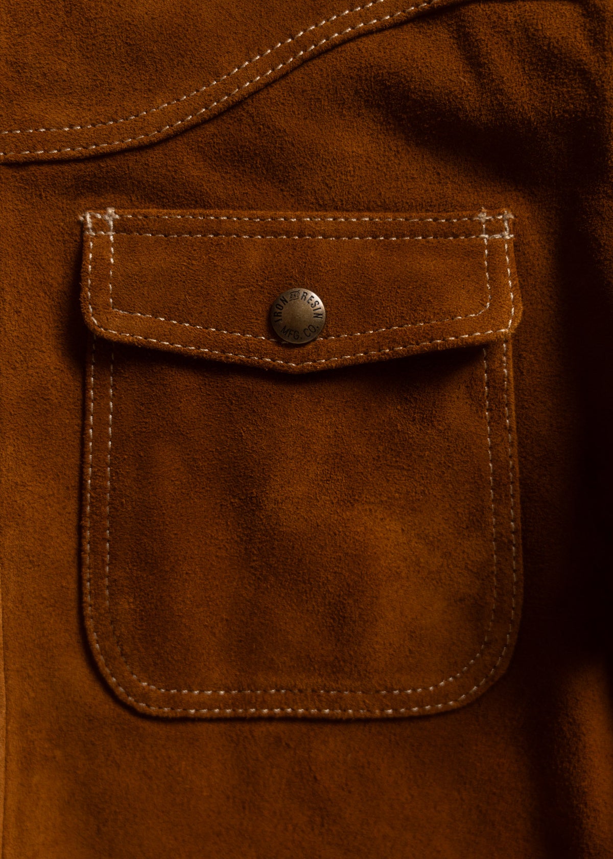 Close-up of a brown suede jacket pocket with a metal button, showcasing detailed stitching and texture.