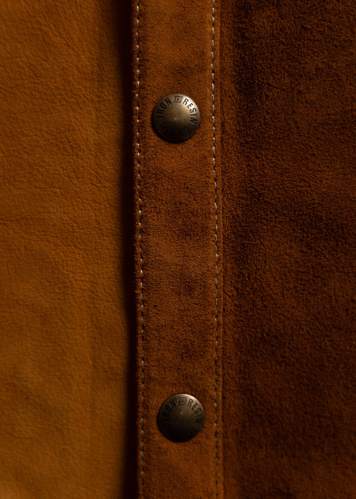 Close-up of brown suede fabric with two metallic buttons engraved "Iron and Resin," showcasing texture and craftsmanship.