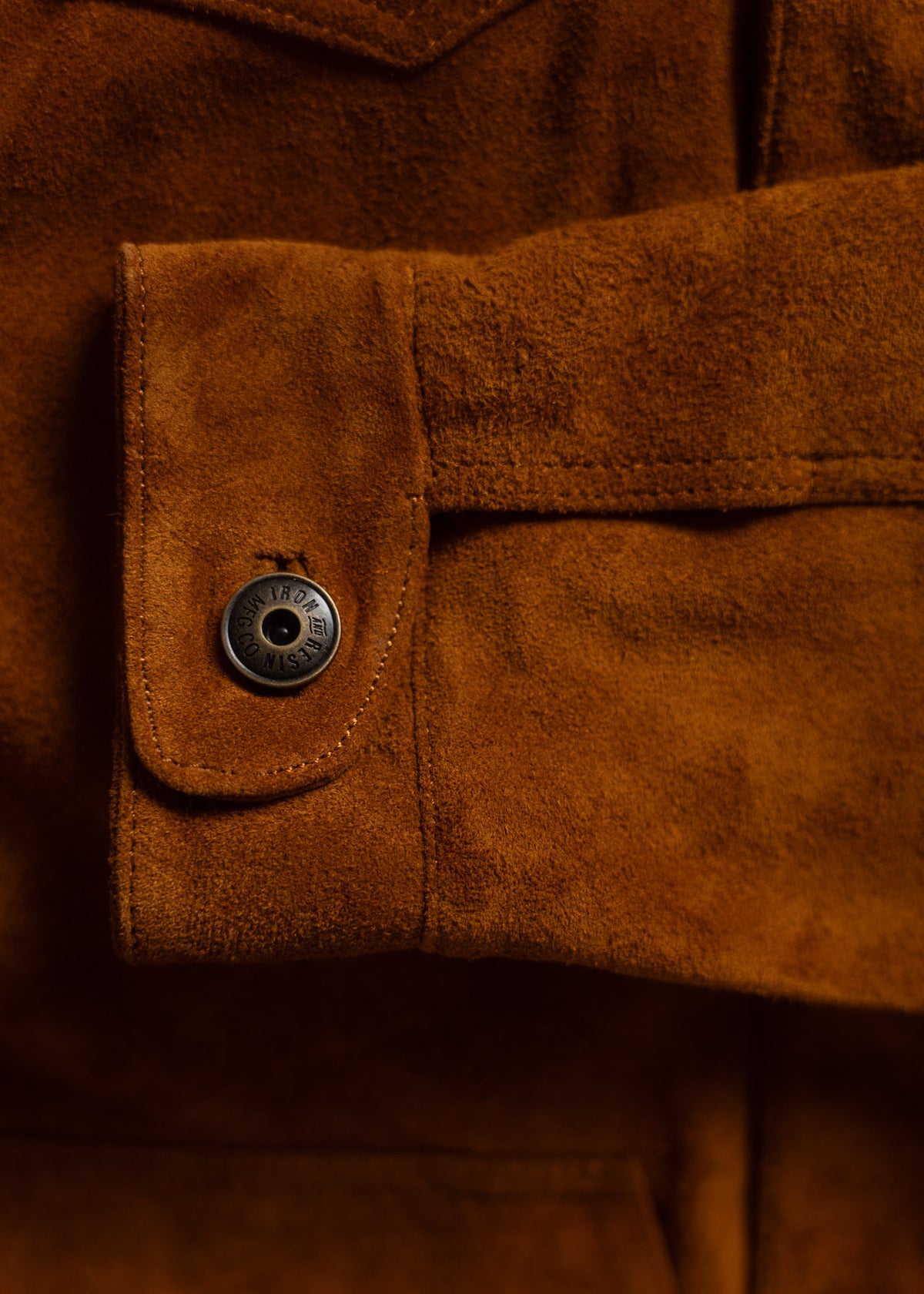 Close-up of a brown suede jacket sleeve with a metal button, showcasing texture and detail.