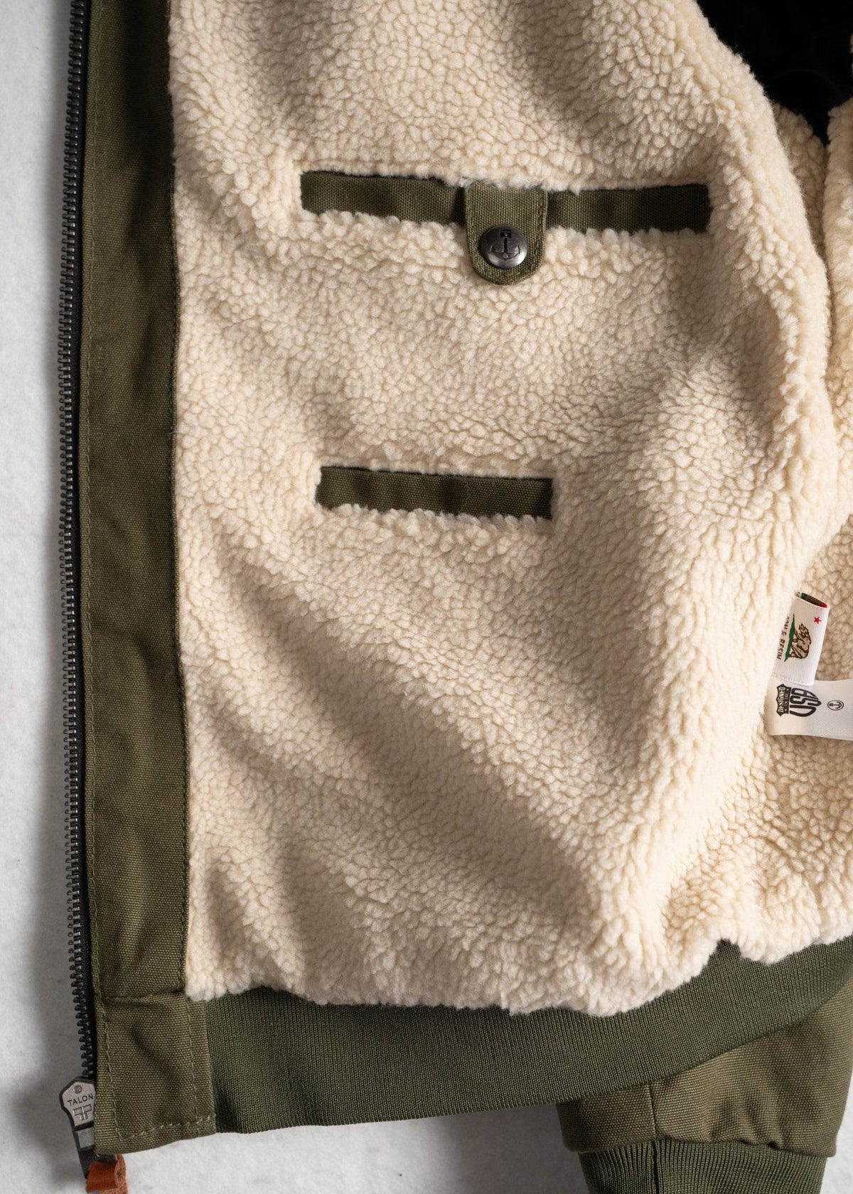 Olive green jacket with soft fleece lining, featuring two pockets and a button detail, lying on a light surface.