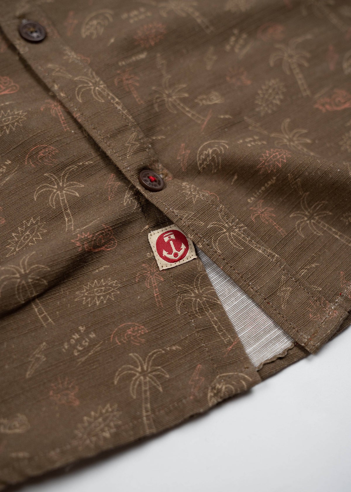 Brown shirt with palm tree pattern, close-up of buttons and red anchor tag detail, showcasing casual tropical style.