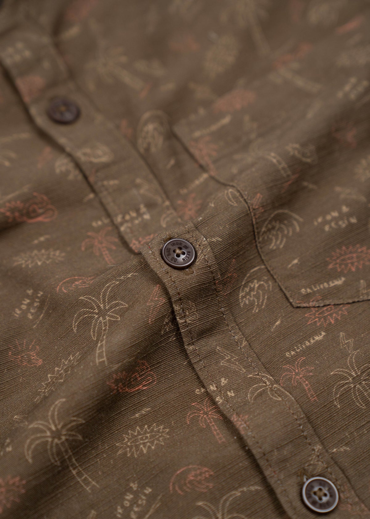 Close-up of a brown shirt with palm tree and text patterns, featuring dark buttons and a casual, tropical design.
