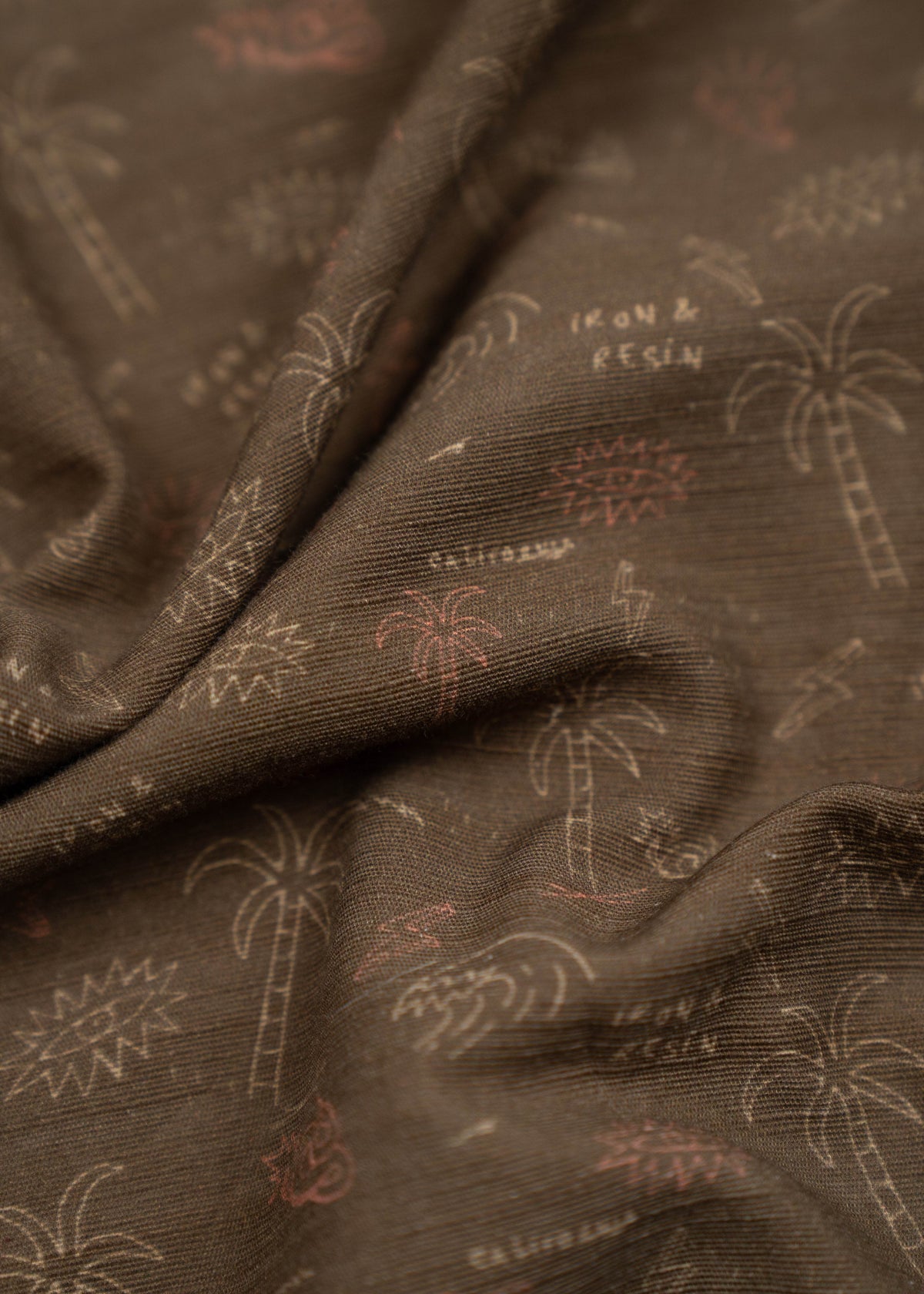 Brown fabric with palm tree and wave patterns, featuring "Iron & Resin" text. The texture is soft and slightly wrinkled.