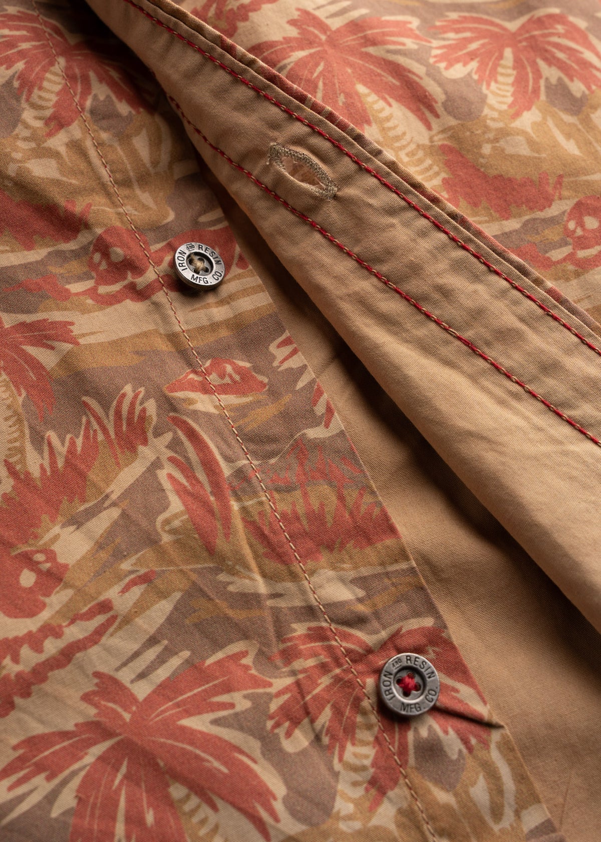 Close-up of a shirt with red palm tree print, red stitching, and visible buttons. Earthy tones create a tropical feel.