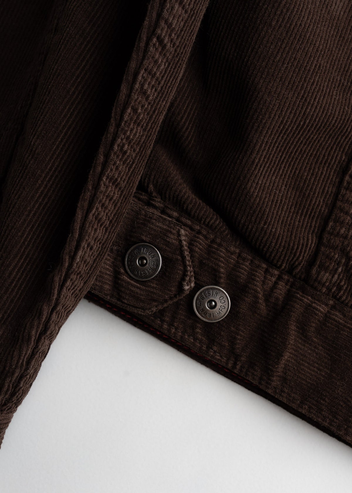 Close-up of brown corduroy fabric with two metal buttons, showcasing texture and detail on a light background.