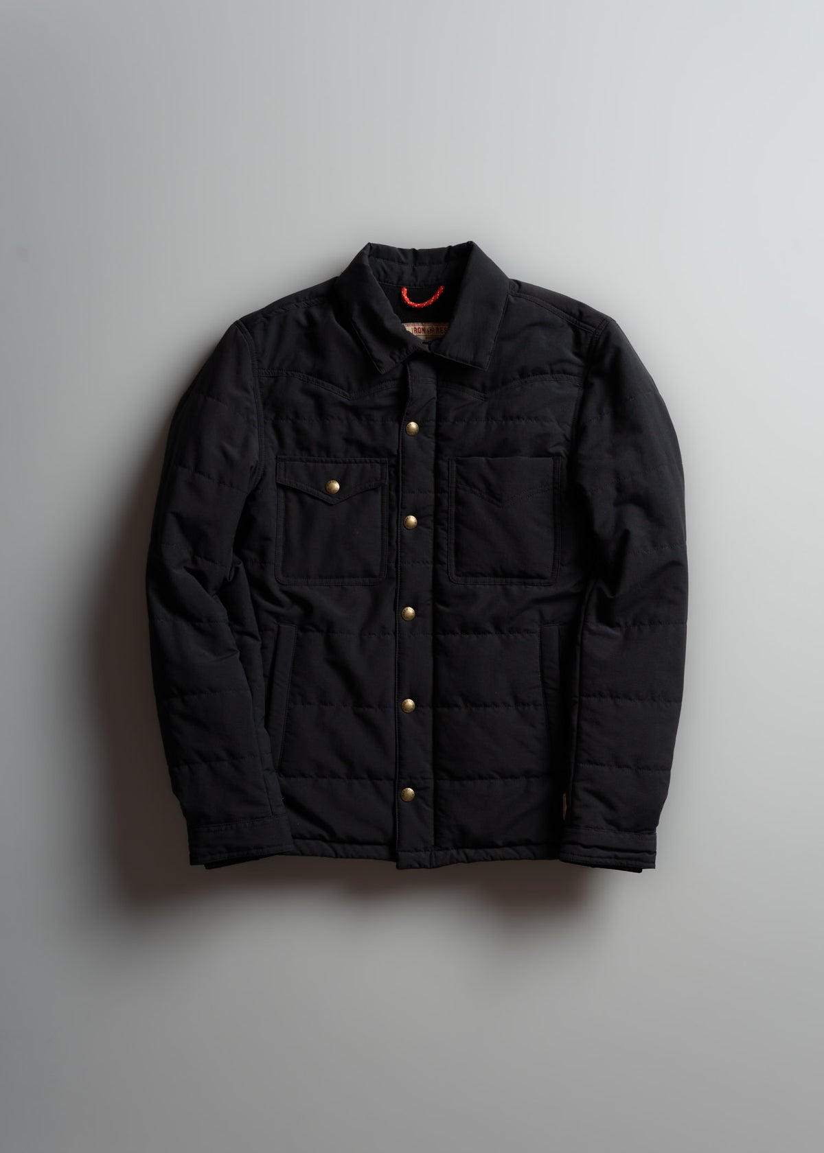 Black quilted jacket with gold buttons and chest pockets, displayed against a light gray background.