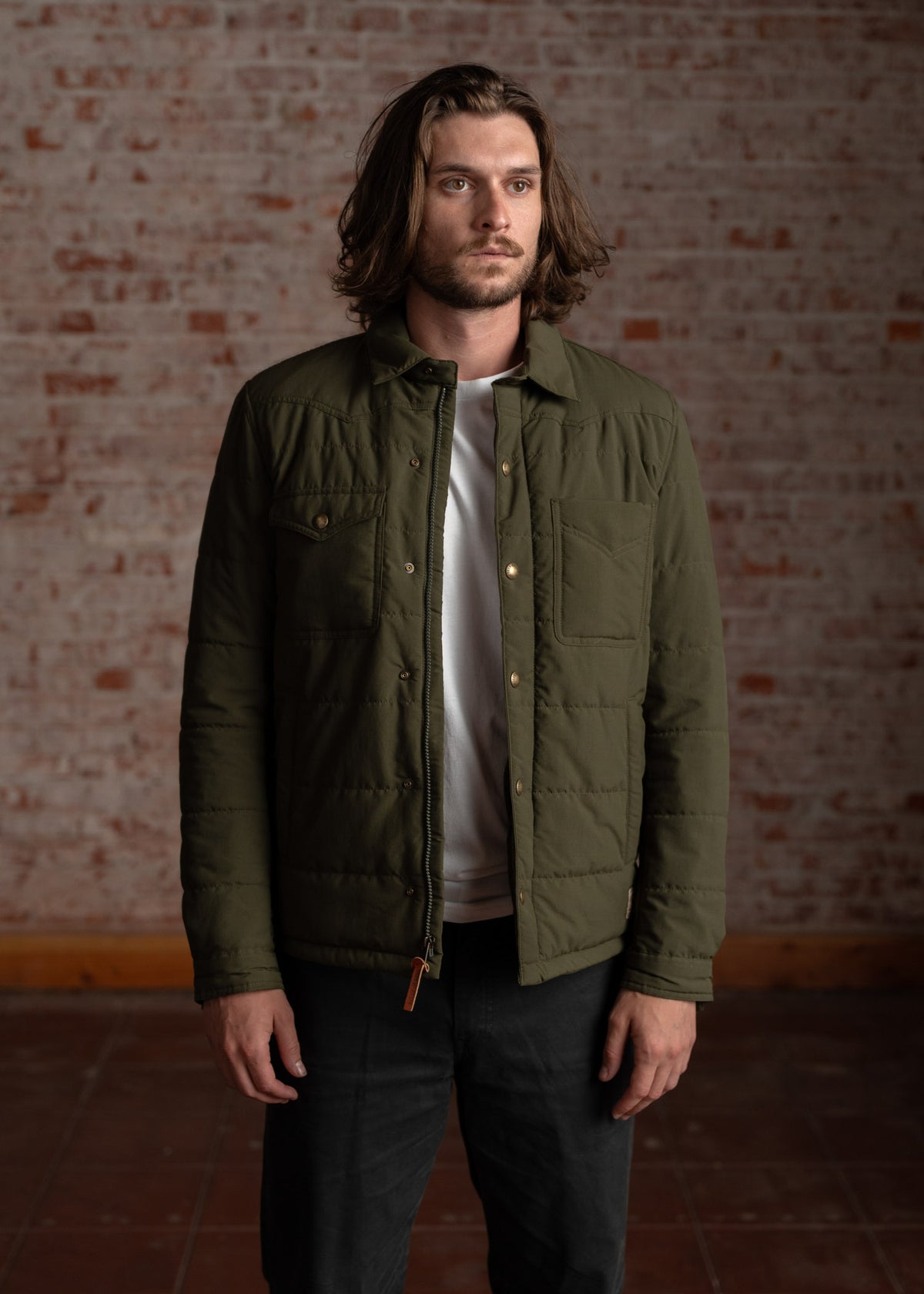 Man in green jacket stands against a brick wall, looking forward. He wears a white shirt and dark pants, exuding a casual style.