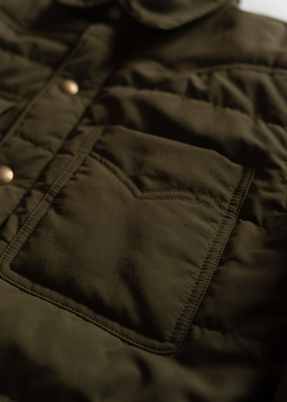 Close-up of a dark green quilted jacket with a front pocket and brass buttons, highlighting its texture and stitching details.