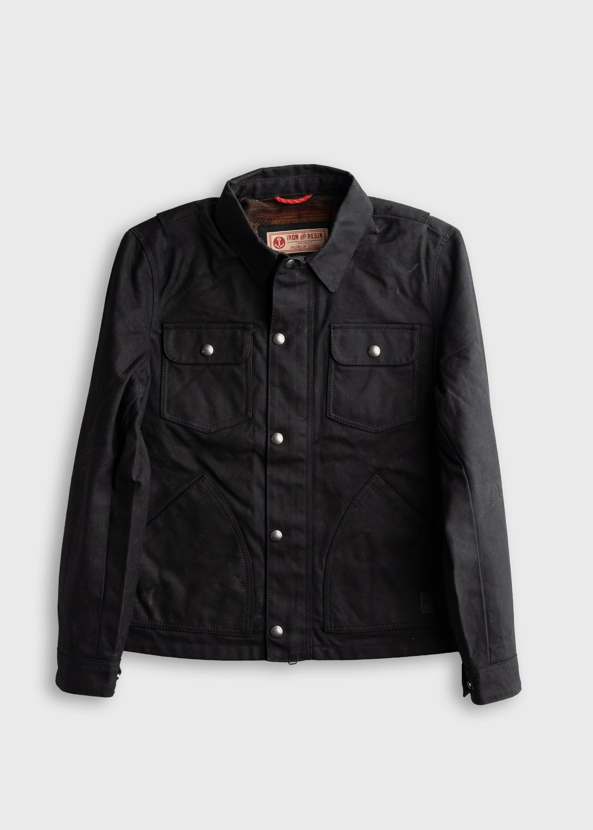 Black waxed canvas jacket with silver buttons and two chest pockets, displayed on a white background.