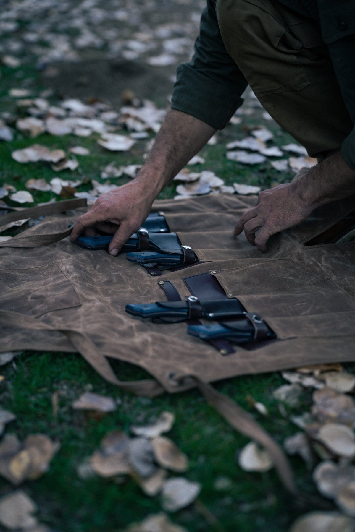 A person arranges knives on a brown canvas mat outdoors, surrounded by fallen leaves on the grass, preparing for a task.