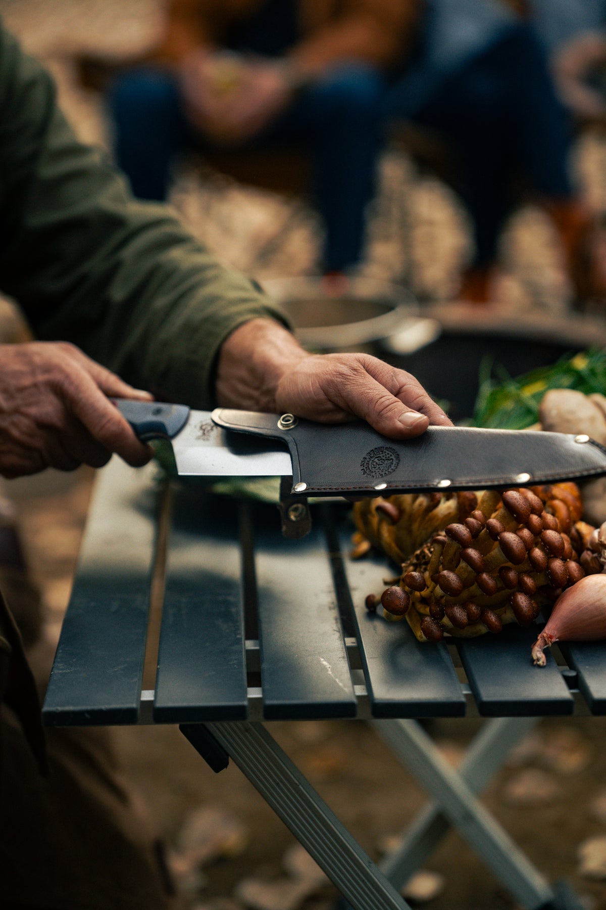A person in a green jacket slices mushrooms on a folding table outdoors, with blurred figures in the background.