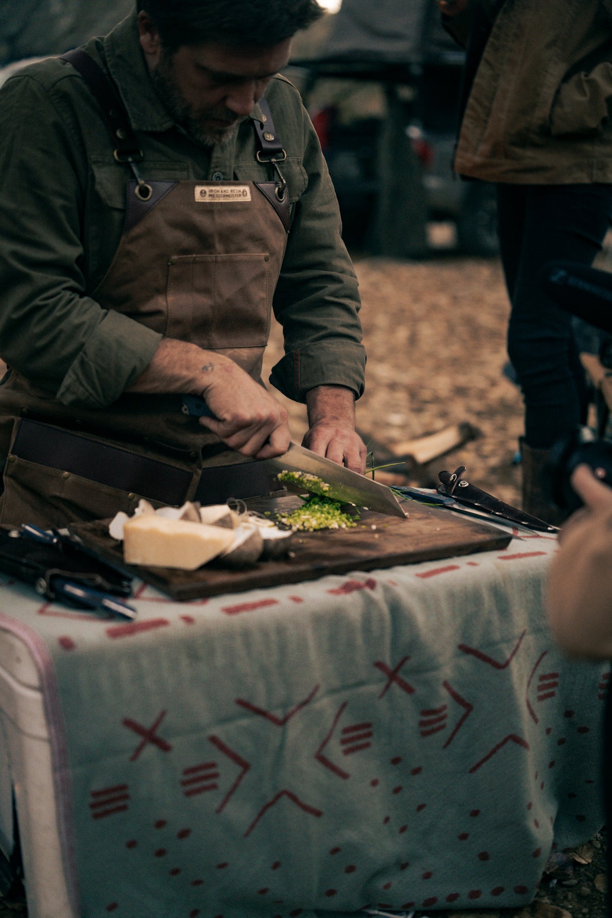 Man in brown apron chopping vegetables on a wooden board outdoors, with cheese and tools on a patterned cloth-covered table.