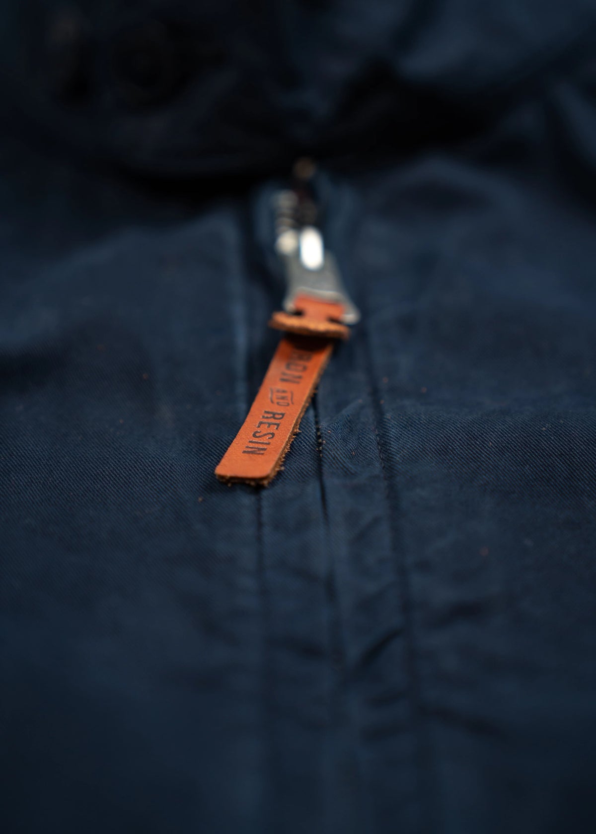 Close-up of a navy jacket with a metal zipper and a brown leather pull tab, featuring embossed text.