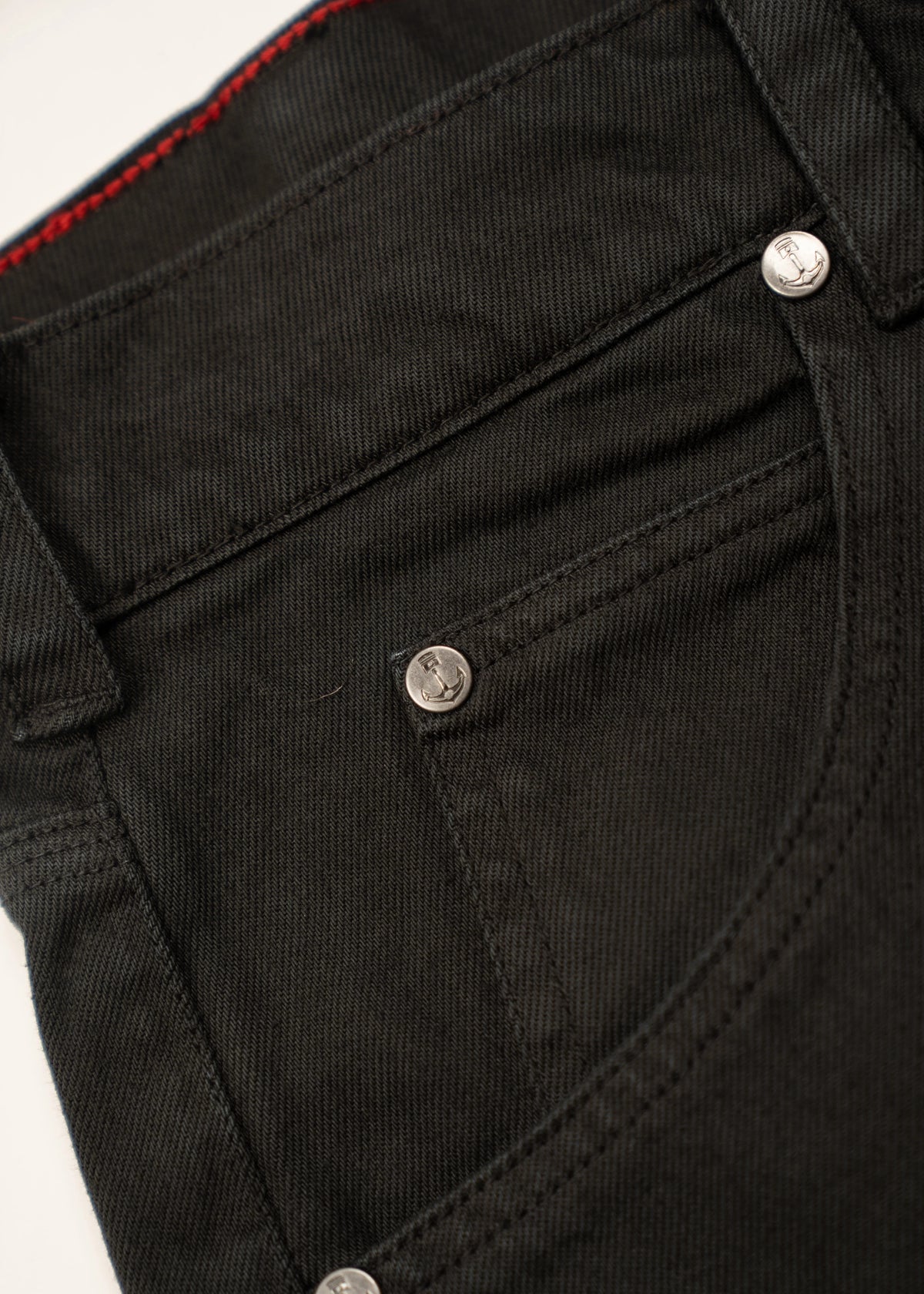 Close-up of black denim jeans pocket with silver rivets and red stitching detail, showcasing texture and craftsmanship.