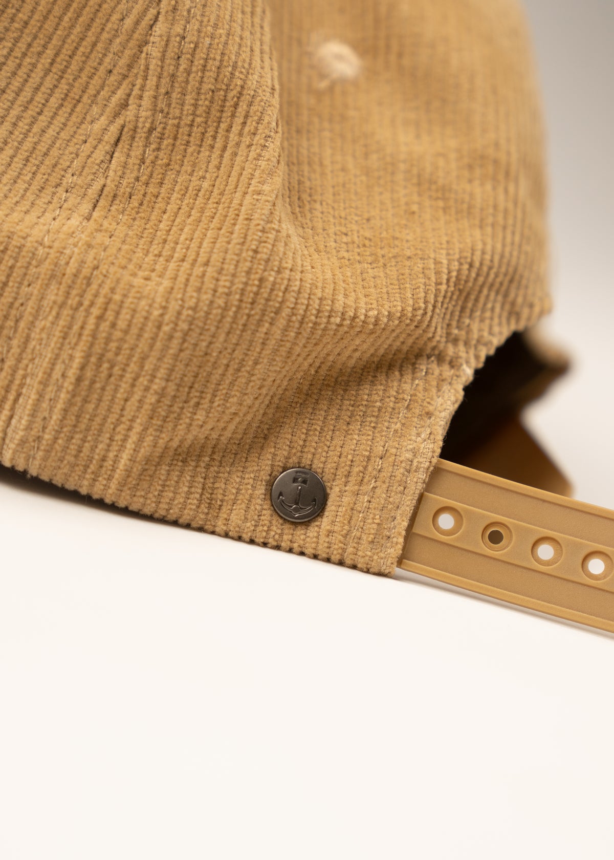 Close-up of a tan corduroy cap with an anchor emblem on the side and an adjustable strap, set against a neutral background.