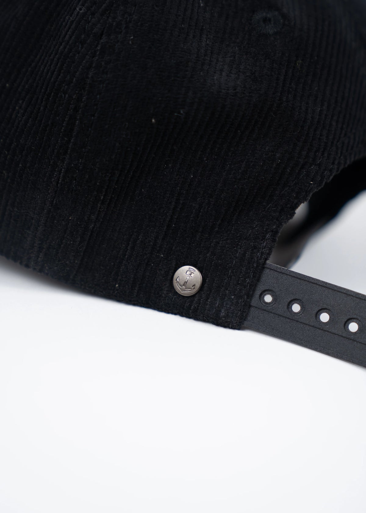 Close-up of a black corduroy cap with an anchor-embossed metal button and adjustable strap on a white background.