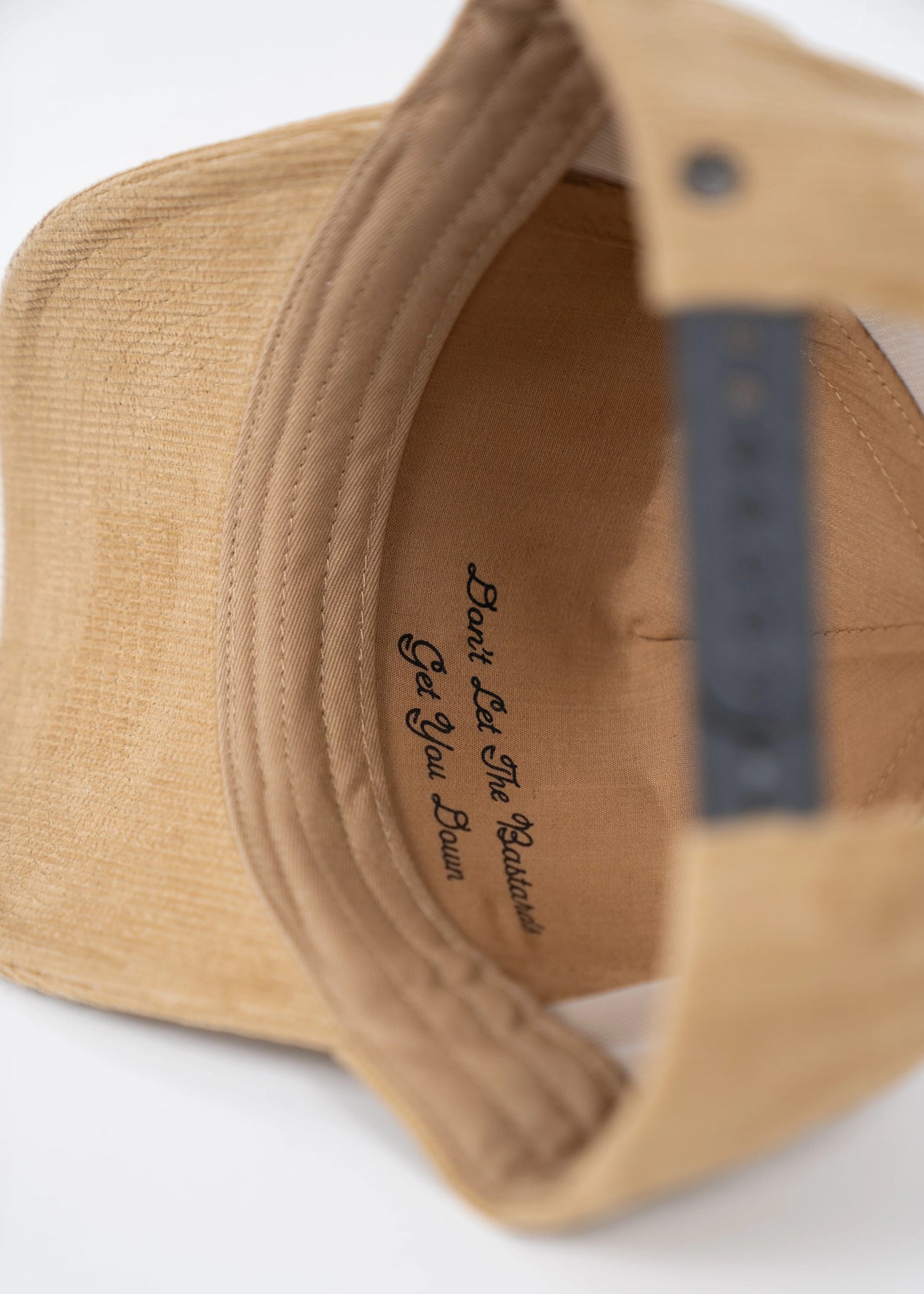 Inside view of a tan cap with adjustable strap, showing the phrase "Don't Let The Bastards Get You Down" printed on the lining.