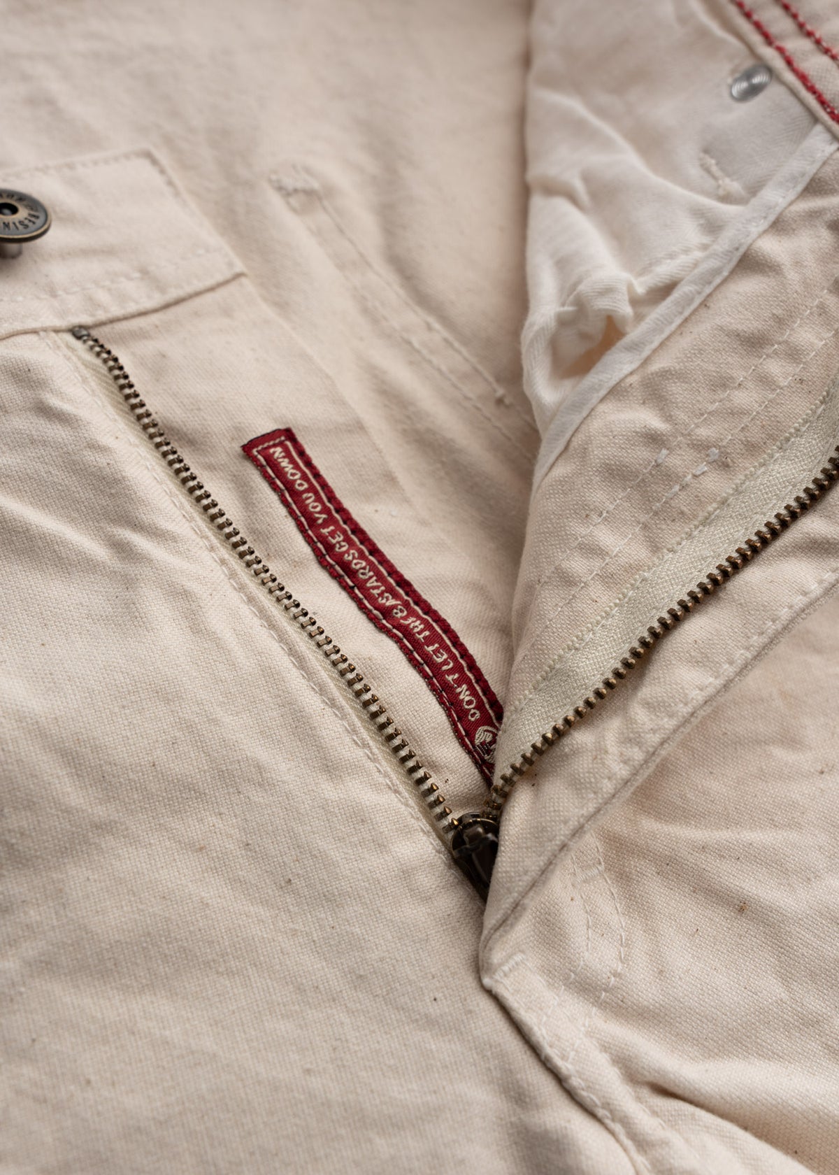 Close-up of beige pants with a visible zipper and a red label reading "DON'T LET THE BASTARDS GRIND YOU DOWN" inside.