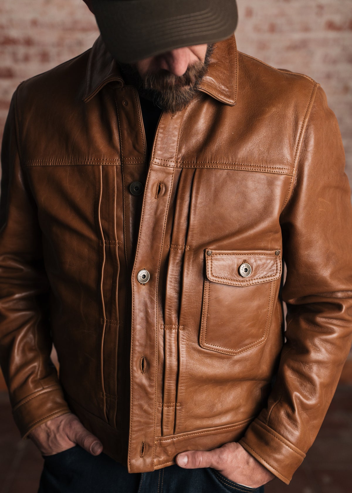 Man in a brown leather jacket and cap stands with hands in pockets, facing down, against a blurred brick wall background.