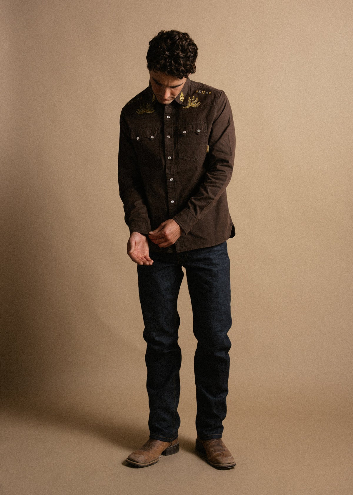 Man in brown embroidered shirt and jeans stands against a beige background, looking down, hands adjusting cuffs, wearing boots.