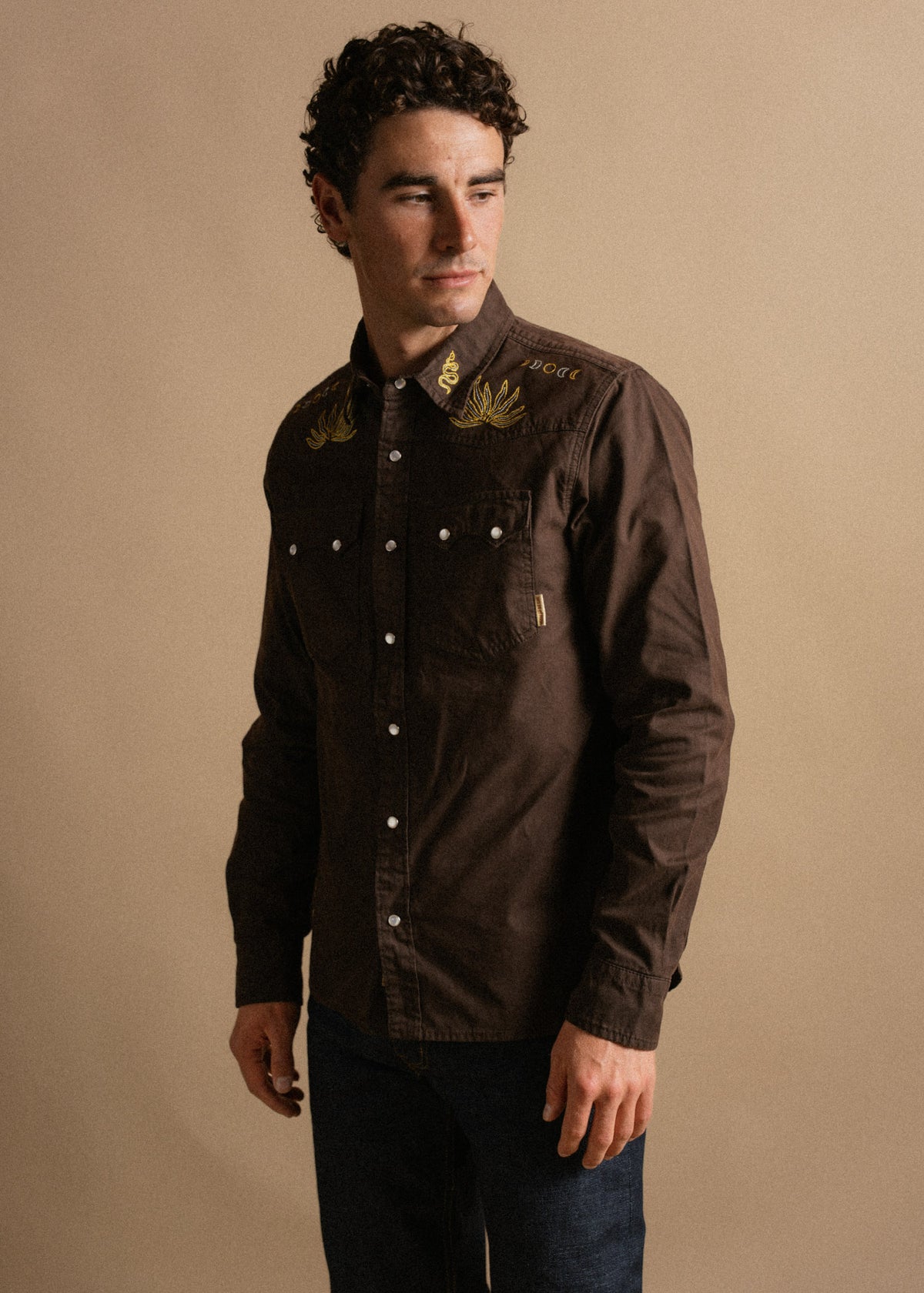 Man in brown embroidered shirt with gold designs, standing against a beige background, looking to the side, hands relaxed.