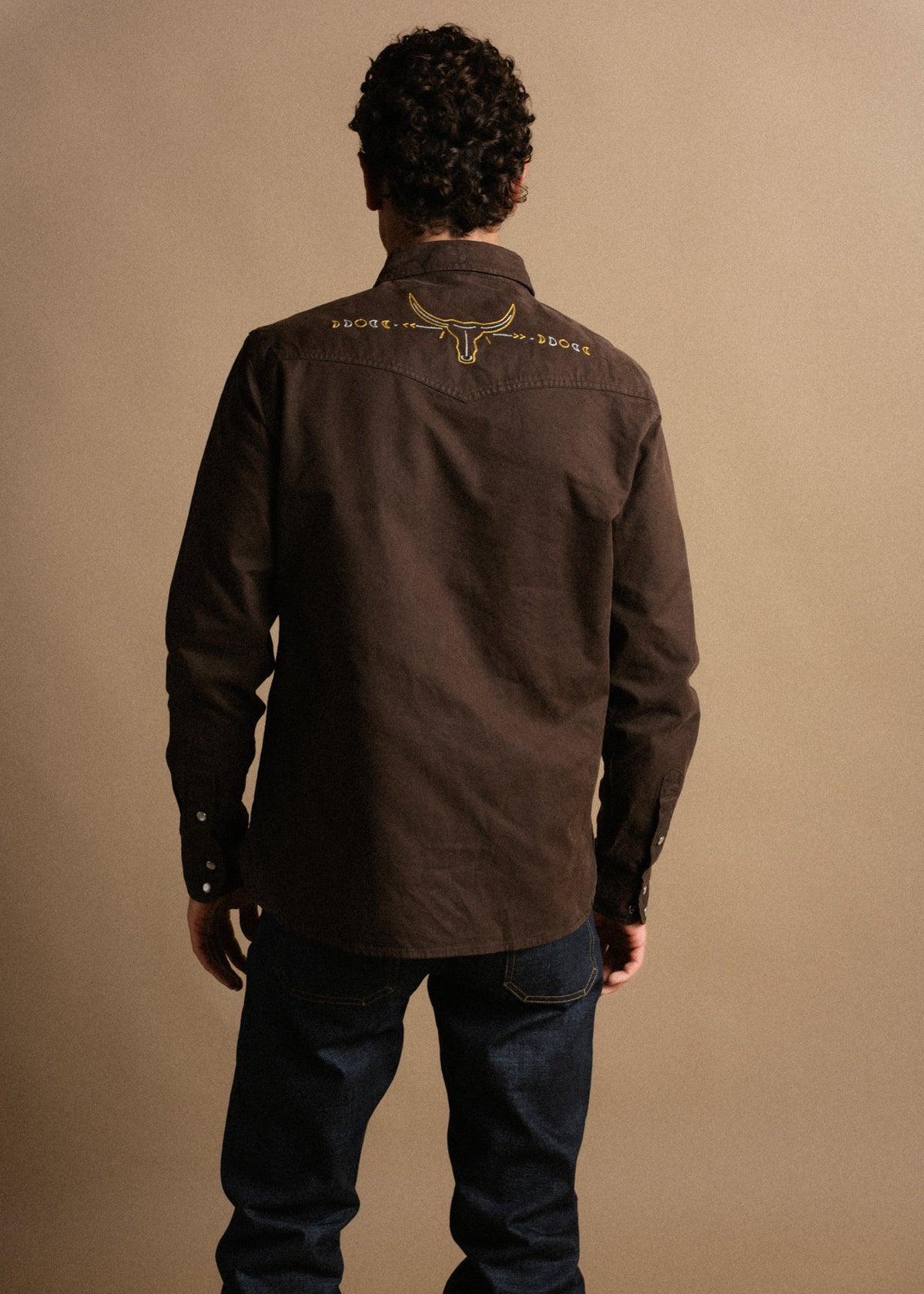 Man in dark shirt with bull embroidery stands against a beige background, facing away. His hands rest by his sides.