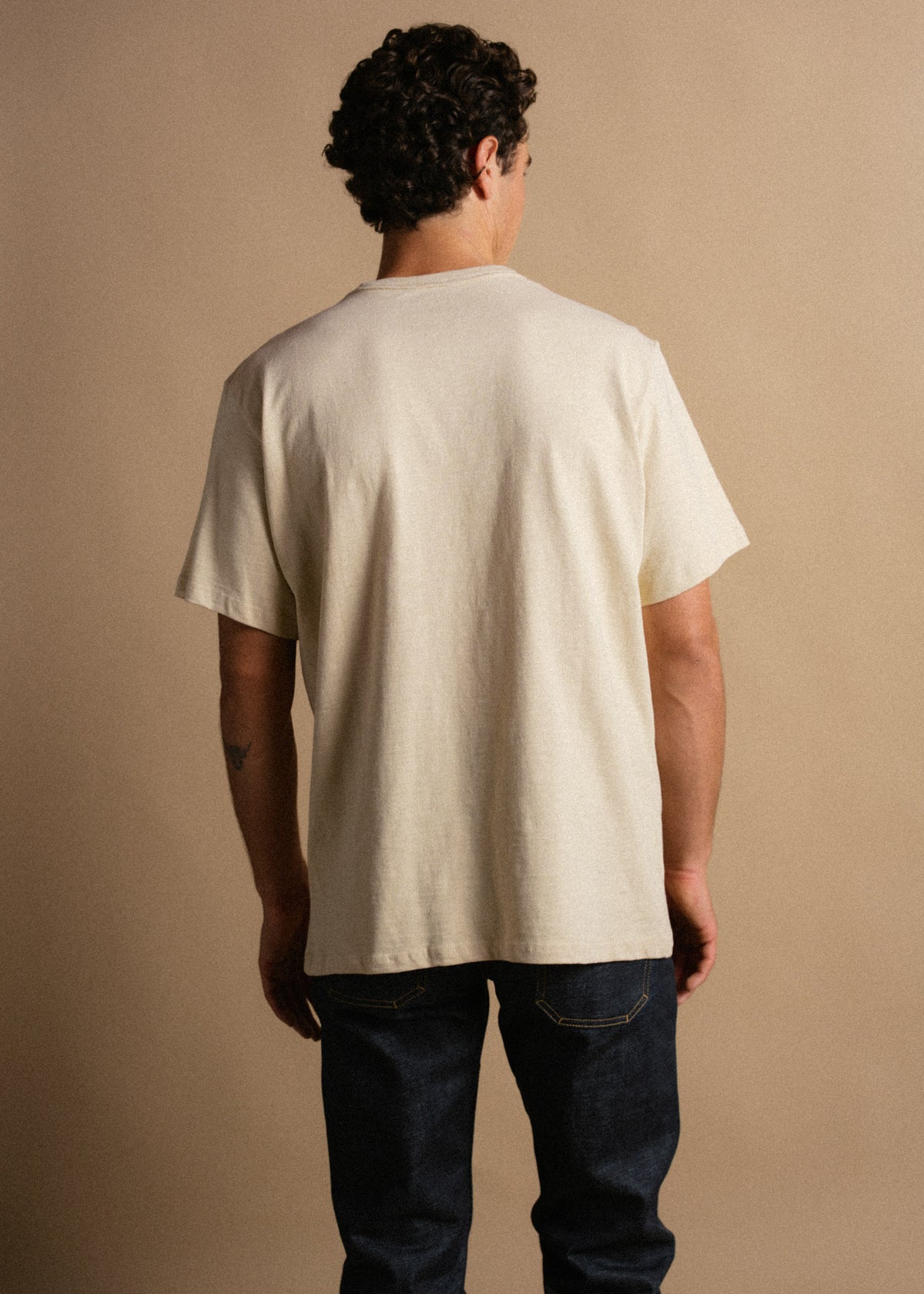 Man in a beige T-shirt and dark jeans stands against a neutral background, facing away. Simple, casual style.