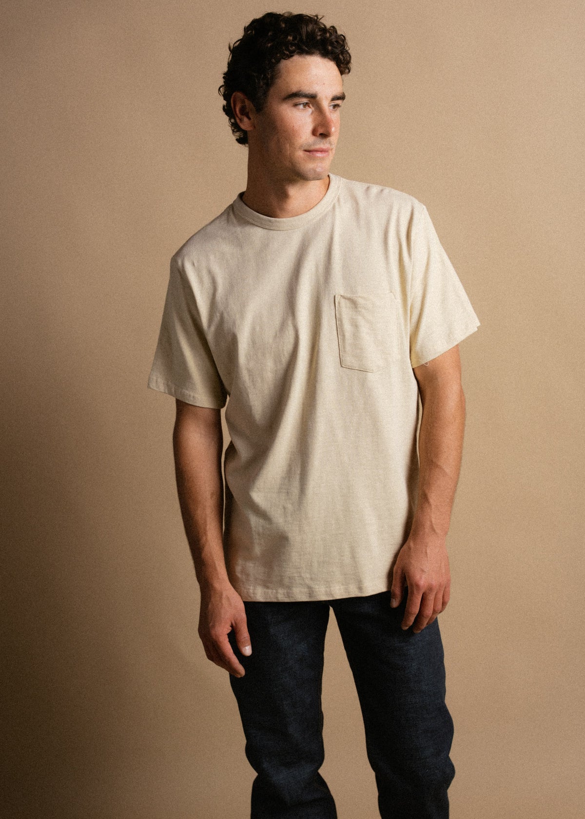Man in beige t-shirt and dark jeans stands against a tan background, looking to the side with a relaxed expression.