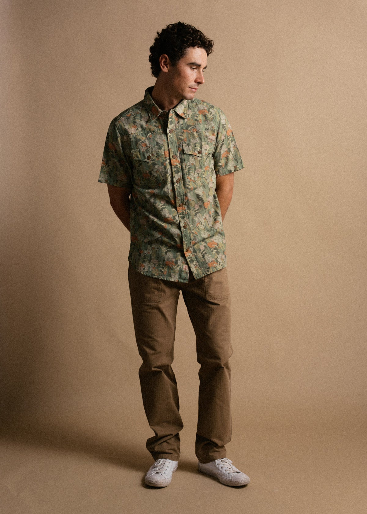 Man in a green patterned shirt and brown pants stands against a beige background, looking to the side with hands in pockets.