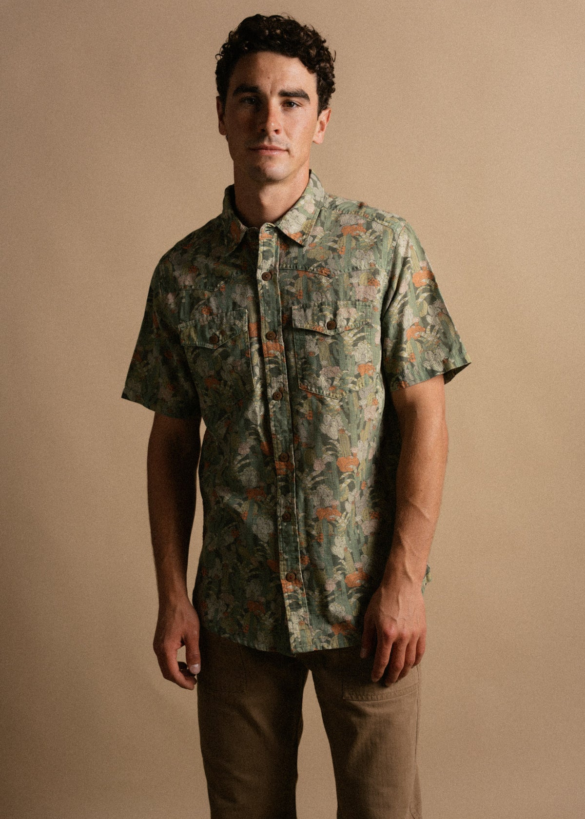 Man in a green floral shirt stands against a beige background, hands relaxed, exuding a casual yet confident demeanor.